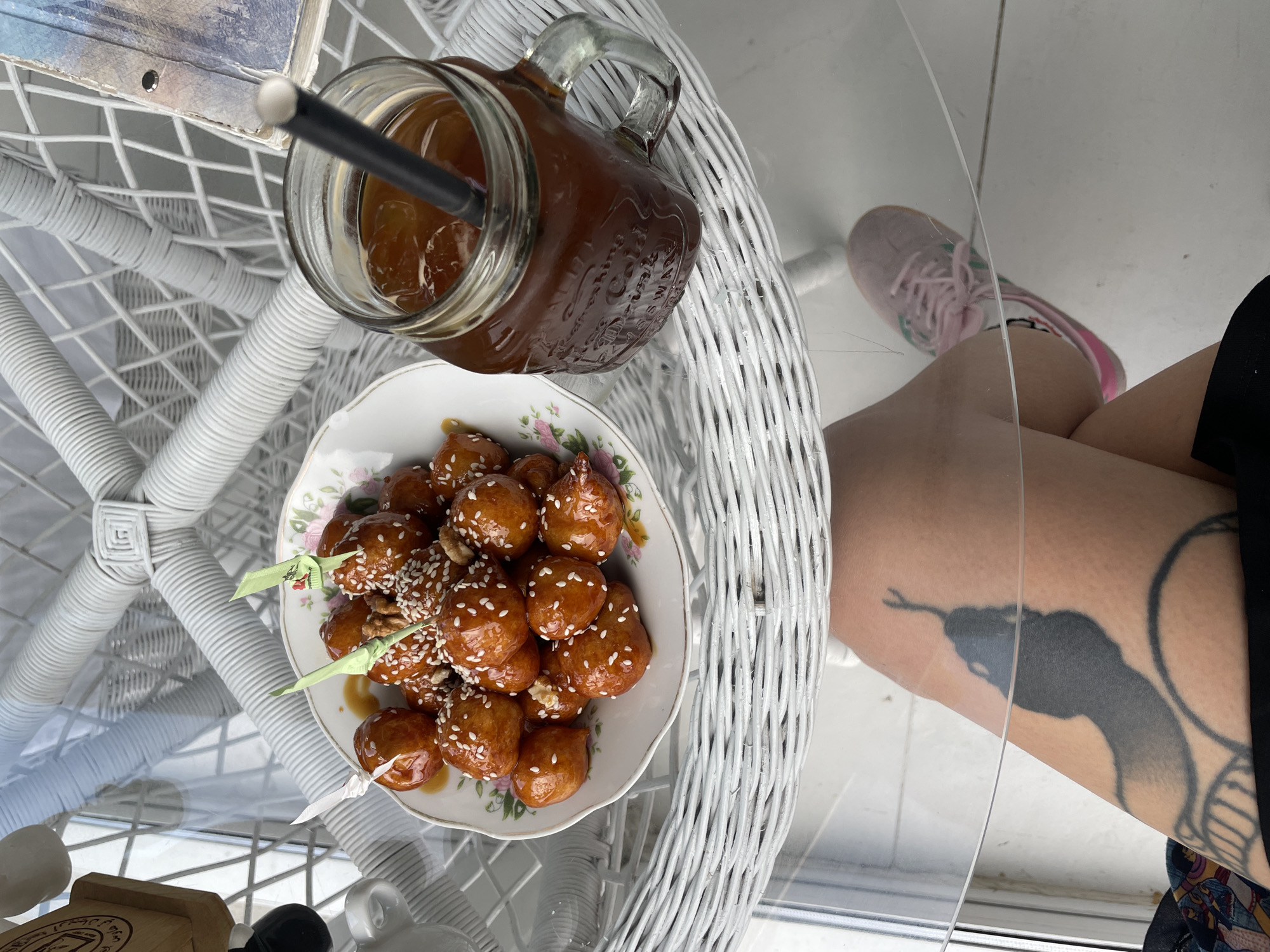 The scene shows a glass-topped wicker table. On the table are a jar of dark liquid with a straw, and a plate of loukoumades (honey doughnuts) topped with sesame seeds. A person is seated at the table; a portion of their leg is visible, including a pink sneaker and a tattoo of a snake. The location is Podgorica, Montenegro, evidenced by a wooden item on the table with Cyrillic text indicating the city.