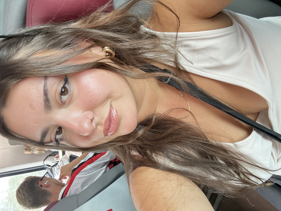 This image depicts a selfie taken by a female individual inside a public transportation vehicle, likely a train, in the vicinity of Castelfranco Emilia, Italy. The primary subject, a young adult female, occupies the foreground, facing the camera with a subtle smile. She presents with light skin, long brown hair, and dark eyes, enhanced by dark eyeliner and shiny, light pink lip gloss. She wears a small silver nose ring on her left nostril and gold hoop earrings. A delicate gold chain necklace is visible around her neck, above a light-colored tank top. A black strap, possibly from a bag, crosses diagonally over her chest. In the mid-ground, to the left, a male individual is partially visible from the back-left shoulder. He has short light-colored hair, wears white wireless earbuds, and a white shirt with red and black striped sleeves, appearing to look downwards. The environment consists of grey passenger seating with red headrests, and a window on the left reveals blurred green vegetation, indicative of a moving conveyance.