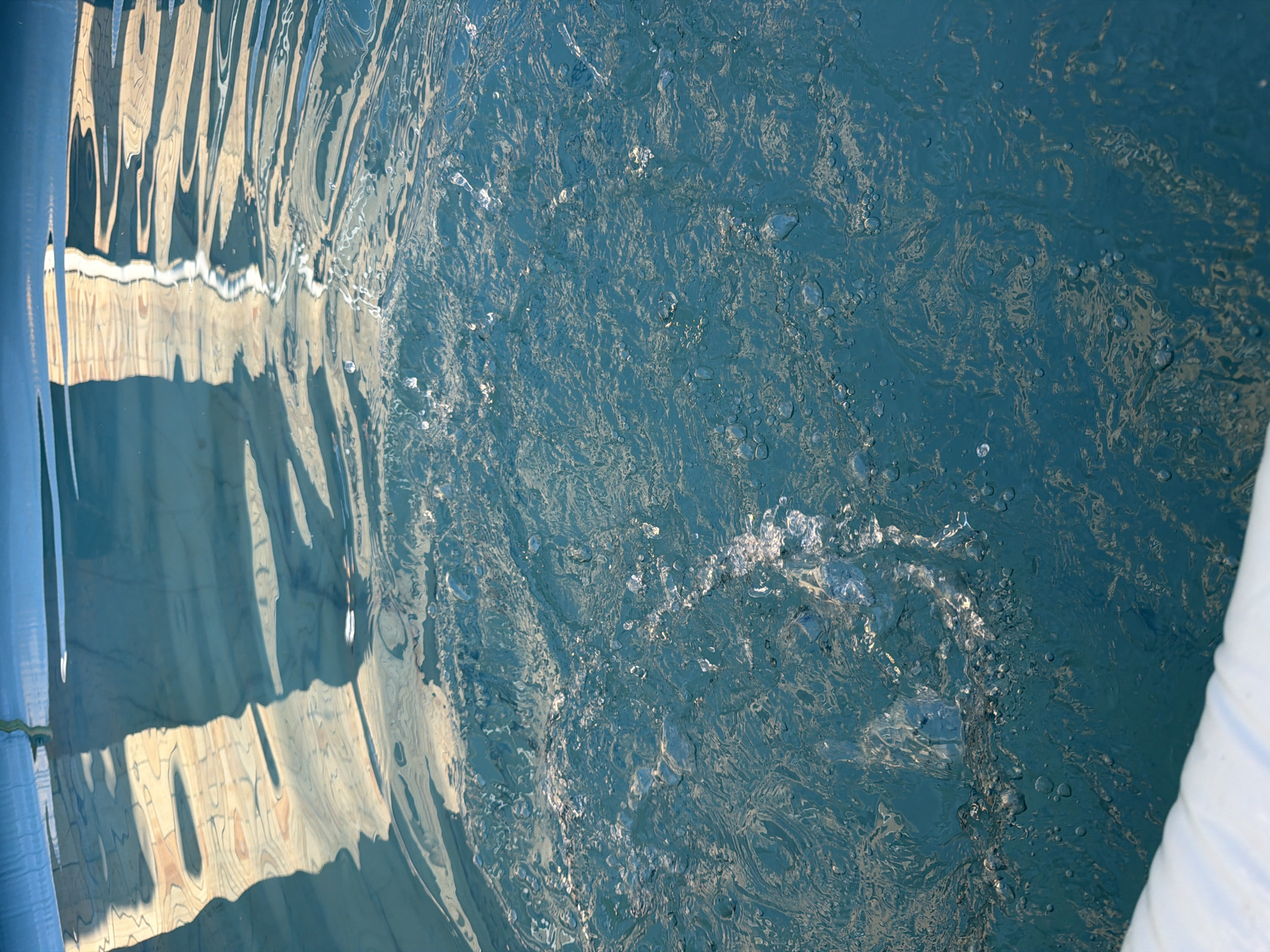 The image shows a close-up, angled view of the surface of a blue swimming pool. The water is disturbed, with ripples, splashes, and bubbles visible. Light reflects off the water's surface, creating abstract patterns of light and shadow. In the upper portion of the image, the reflections of what appear to be vertical structures with an overhead canopy are distorted by the movement of the water. The lower left corner of the image shows a curved, white inflatable edge of the pool. The scene appears to be outdoors during daylight. There are no people or animals visible in the image.