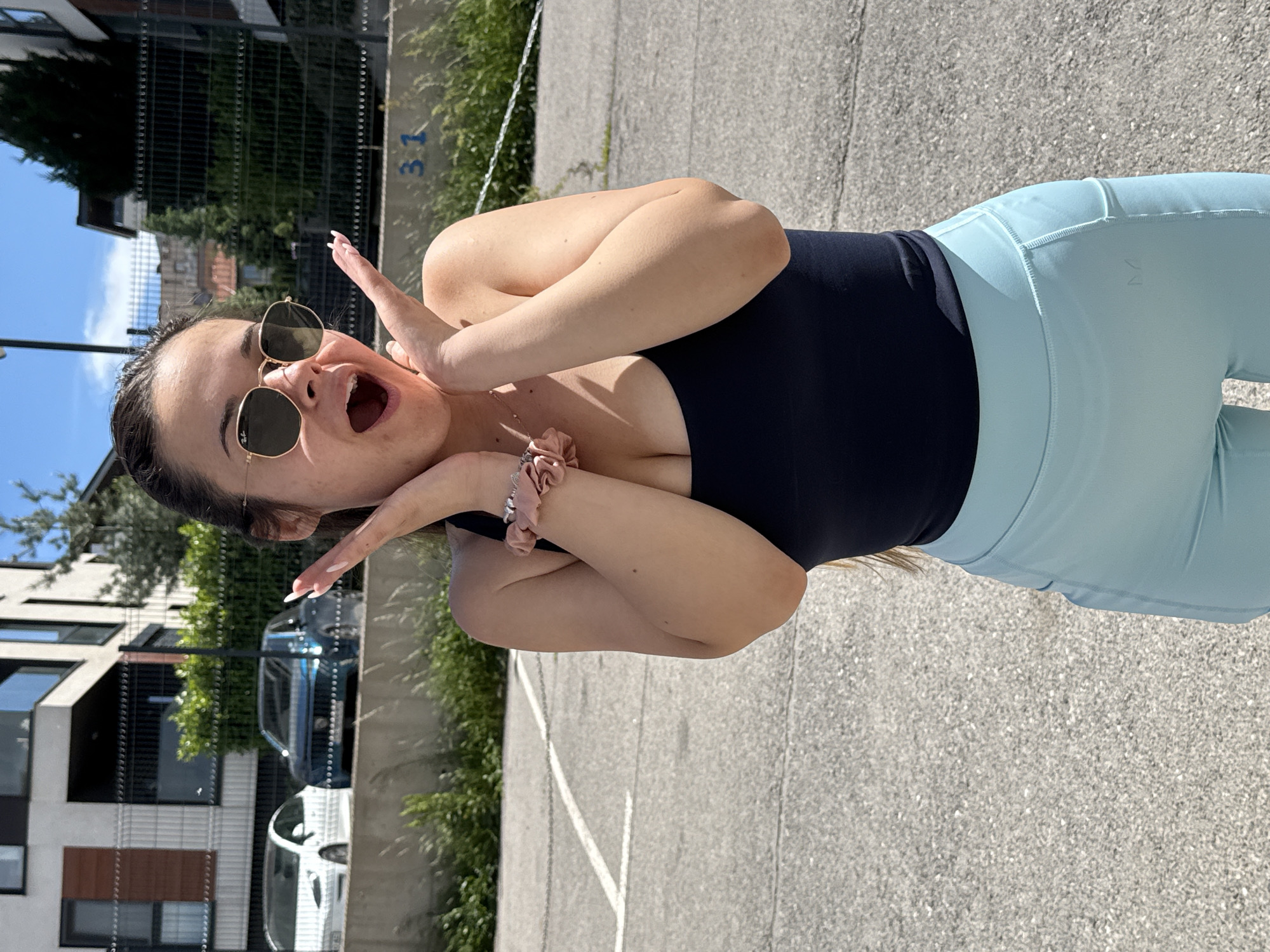 A young woman with brown hair is photographed in a medium shot, outdoors, in what appears to be an urban setting in Sofia, Bulgaria. She is wearing a dark-colored top, light blue leggings, and round sunglasses. She has her hands up to her face with an open-mouthed expression. A scrunchie adorns her wrist. The background shows an asphalt area, a fence with a number "31" visible, green trees and shrubbery, and building structures including cars. The buildings have a modern architectural design. The photo is rotated slightly, causing the vertical elements to appear slanted.