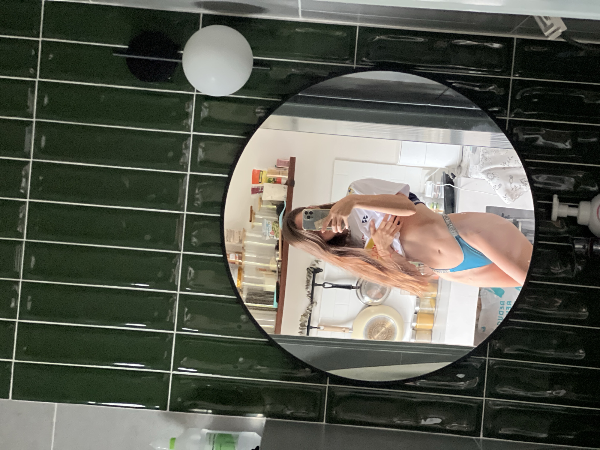 The image is a bathroom selfie taken in an unidentified location in Unknown, Unknown. A woman is positioned in the reflection of a circular mirror, holding a smartphone to capture her image. She is wearing a white top and blue underwear. Behind her in the mirror's reflection, there is a kitchen scene with shelves containing jars and utensils. The bathroom itself features dark green, rectangular tiles on the walls, a light fixture above the mirror, and various bathroom amenities on the countertop below the mirror.