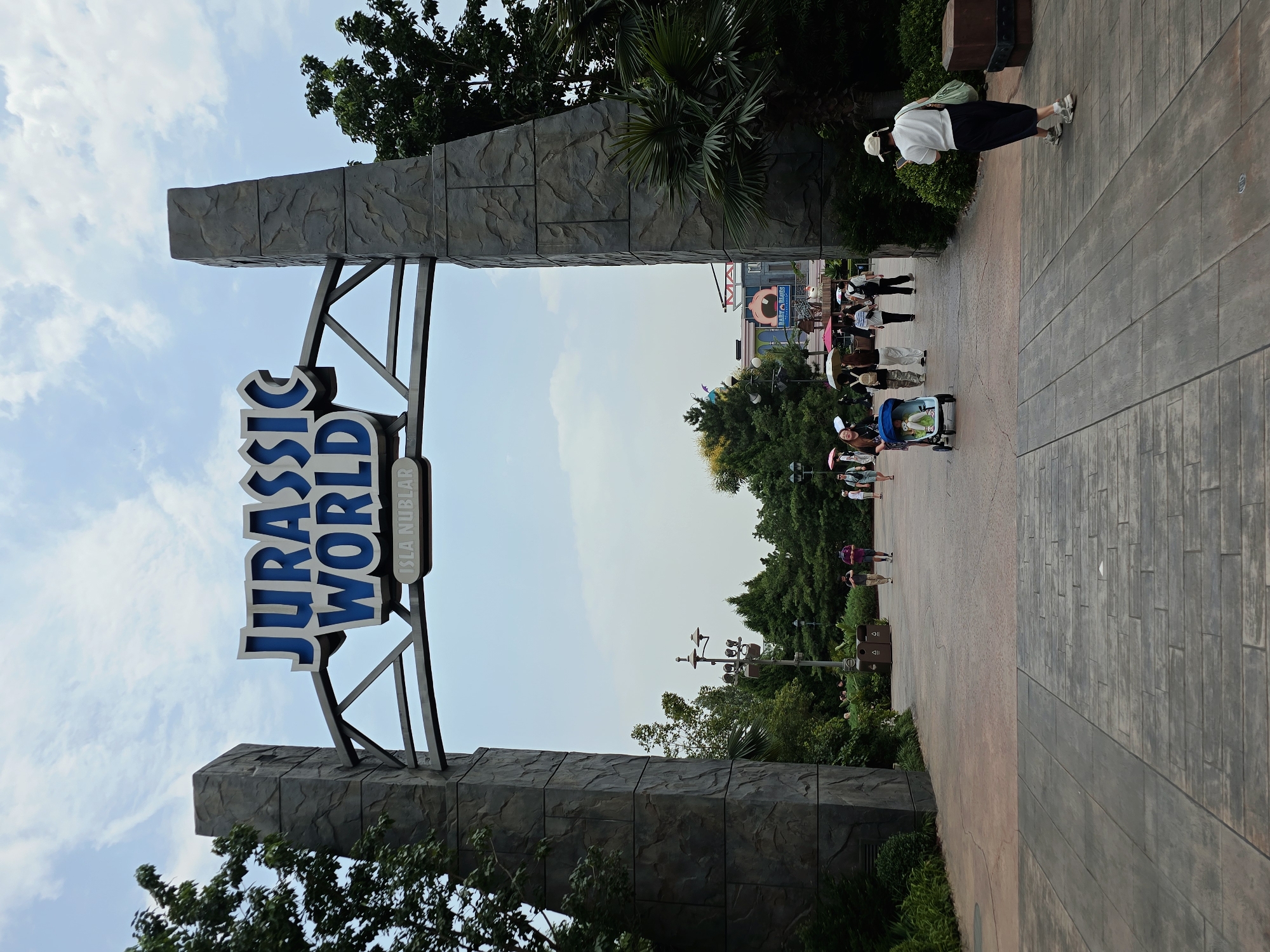The image captures the entrance to Universal Beijing Resort's Jurassic World Adventure Park in Tongzhou District, China. A large, stone-textured archway frames the "JURASSIC WORLD" logo with "ISLA NUBLAR" below it. The pathway leading through the archway is paved with interlocking stone tiles. Several people are walking in the area, some carrying umbrellas. A person in the foreground is walking away from the camera, looking at their phone, and carrying a green bag. In the background, there are lush green trees and buildings with colorful signage, one of which appears to be related to a character with a large mouth. The sky is partly cloudy.
