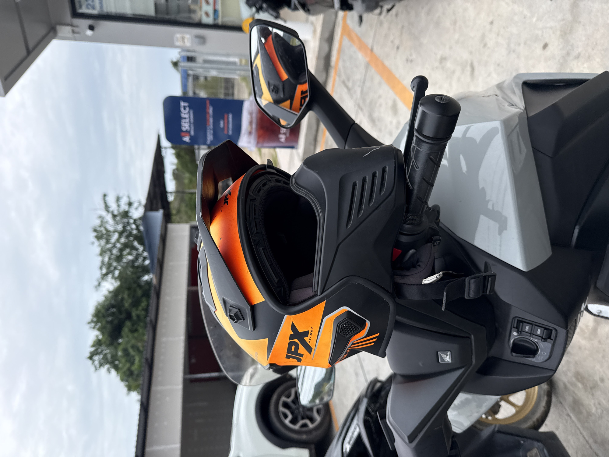A close-up, eye-level outdoor shot captures a JPX helmet with orange and black accents resting on the handlebars of a light gray motorcycle. The helmet is open, revealing its interior padding. The motorcycle's handle grips and part of the dashboard, including ignition and control buttons, are visible. In the background, a white SUV is parked on a concrete surface marked with yellow lines, suggesting a parking lot. Further back, a building with a white overhang and trees are present. A blue sign with "A SELECT" printed on it is also visible, along with a vending machine dispensing a red beverage. The sky is overcast. The scene is located in the Ko Samui District, Thailand.