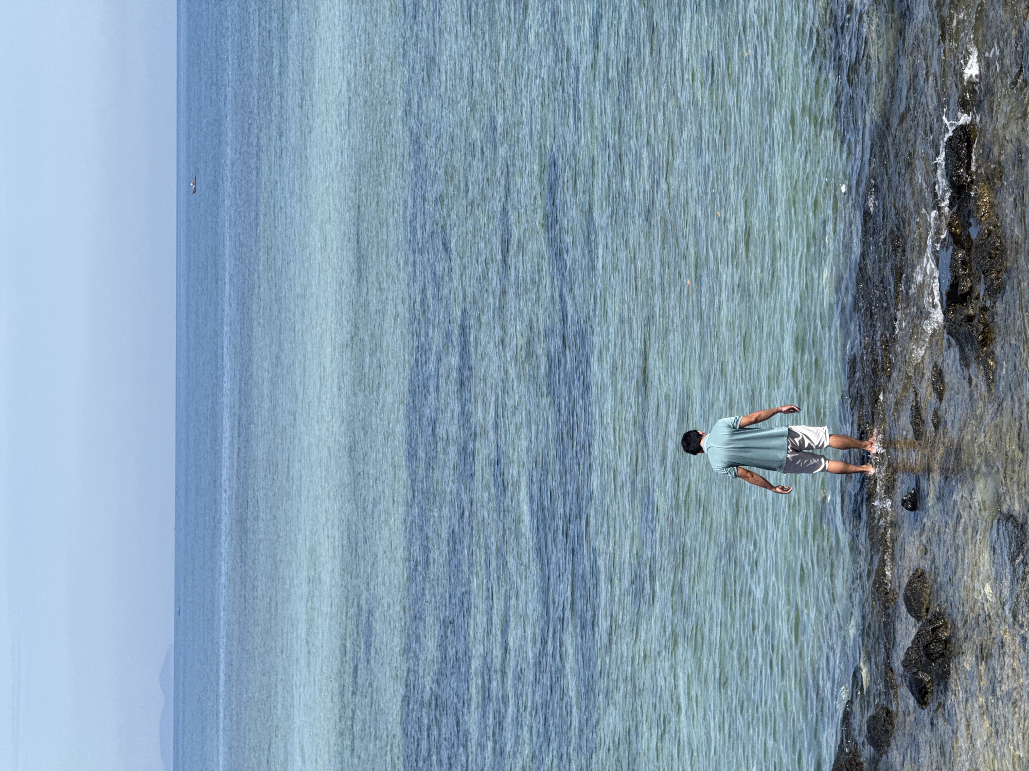 A man stands at the edge of the water, facing away from the camera, on a rocky shore. He is wearing a teal polo shirt and light-colored shorts. His feet are submerged in the clear, shallow water. The sea stretches out before him, a gradient of blues and greens, with a faint outline of land on the horizon. A small boat is visible in the distance. The scene appears to be a tranquil coastal setting.