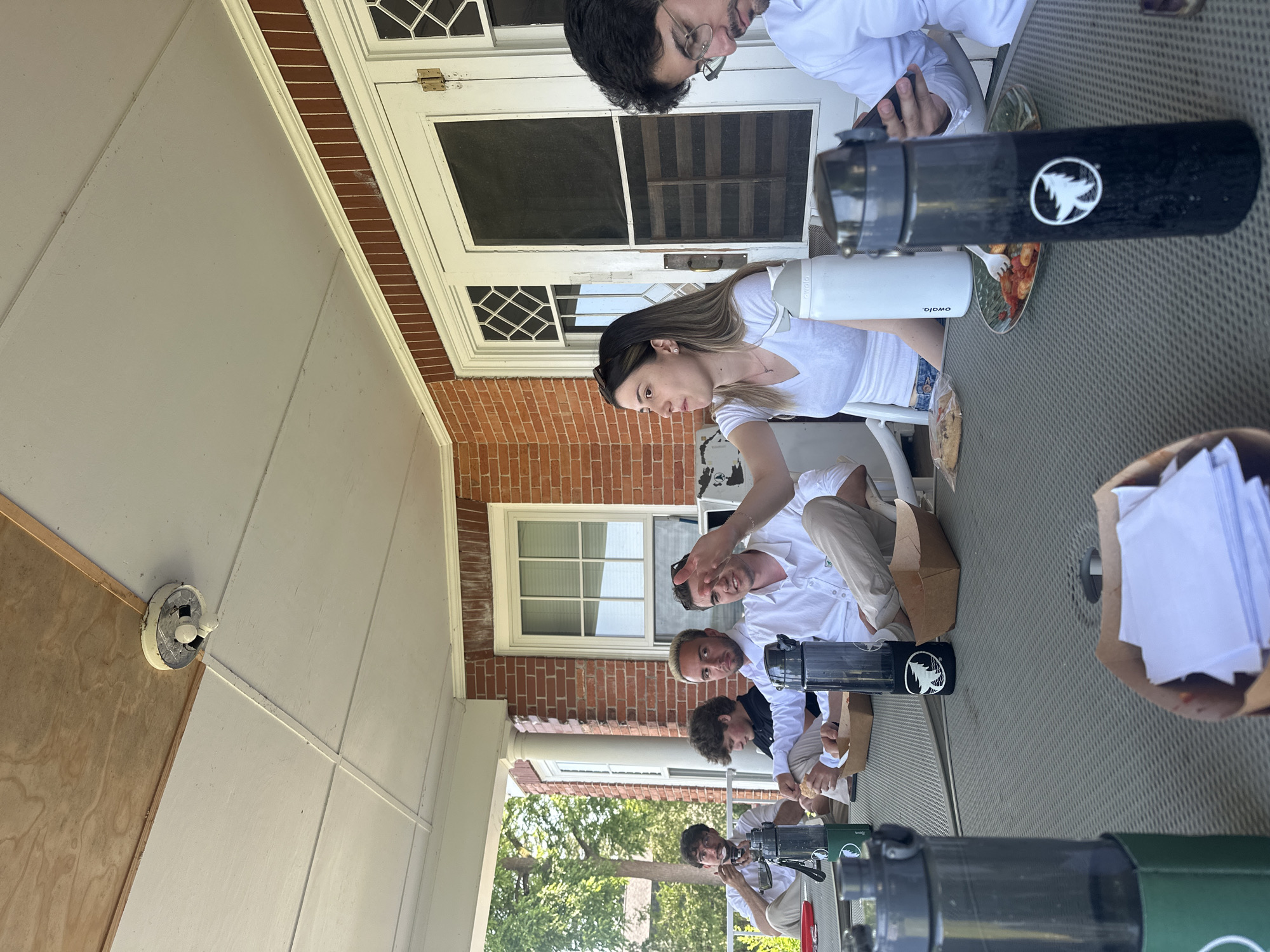 This media file depicts a group of people gathered on a porch, likely during a daytime social event. The scene is outdoors, with a brick building and a white-columned porch visible in the background, suggesting a residential setting. A table with a textured surface occupies the foreground, covered with various items including food, drinks in water bottles, and paper.
The subjects are several individuals, some of whom are wearing white shirts. They appear to be interacting with each other and the objects on the table. One woman in a white t-shirt is looking down, holding a water bottle. A man with glasses and a white shirt is looking towards the camera. Another man is gesturing with his hand while looking at the camera. The overall mood seems relaxed and casual.
Notable details include several reusable water bottles, one black with a white logo of a tree and a bird, and another white with text that reads "owala." There are also brown paper bags, some containing food items and others filled with white paper. The time of day appears to be daylight, and the weather seems pleasant, conducive to an outdoor gathering. The location cues, such as the architecture of the building and the porch, suggest a possibly suburban or historic setting in the United States. The individuals' expressions and interactions suggest a friendly and informal occasion.