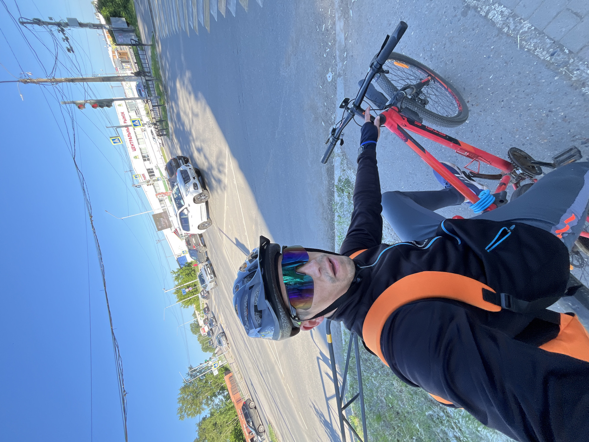 In the city of Chaikovsky, Russia, a man wearing a black cycling jacket, grey pants, a black helmet, and iridescent cycling glasses is seated on a red bicycle. He is positioned on the right side of the frame, with the bicycle's handlebars and front wheel visible. Behind him, a street intersection is visible with a pedestrian crossing. Cars are present on the road, and a building with the Cyrillic text "ЦЕНТРАЛЬНЫЙ РЫНОК" (Central Market) is in the background. The sky is clear and blue. The overall scene depicts an urban environment on a sunny day.