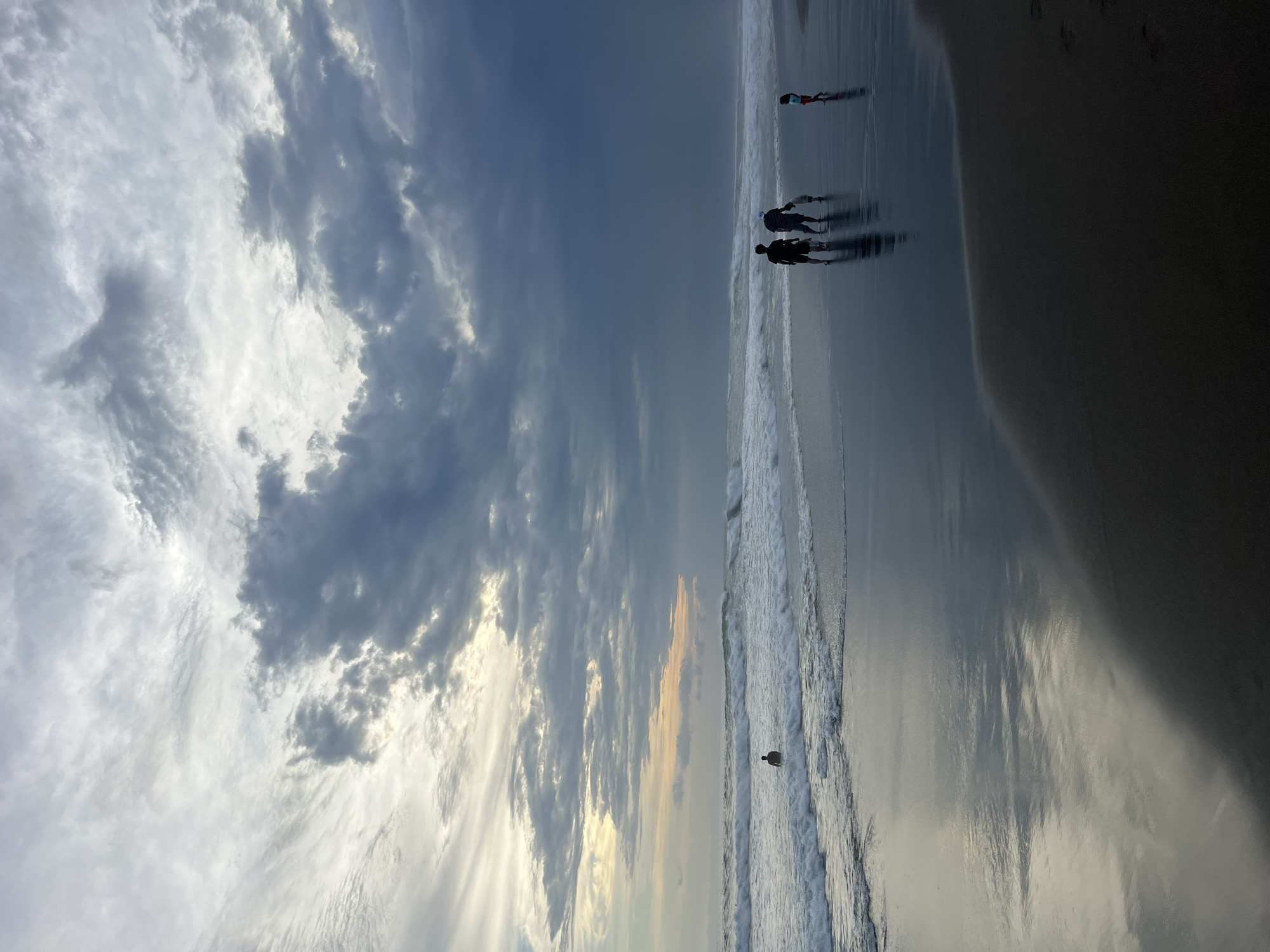 The image shows a beach scene in North Kuta, Indonesia. The photo is rotated 90 degrees counterclockwise. A group of four people are walking along the shoreline at the top of the image. The beach is wet and reflective. A single bird is standing near the water's edge. The sky is filled with clouds, with the lighter portion of the sky near the horizon.