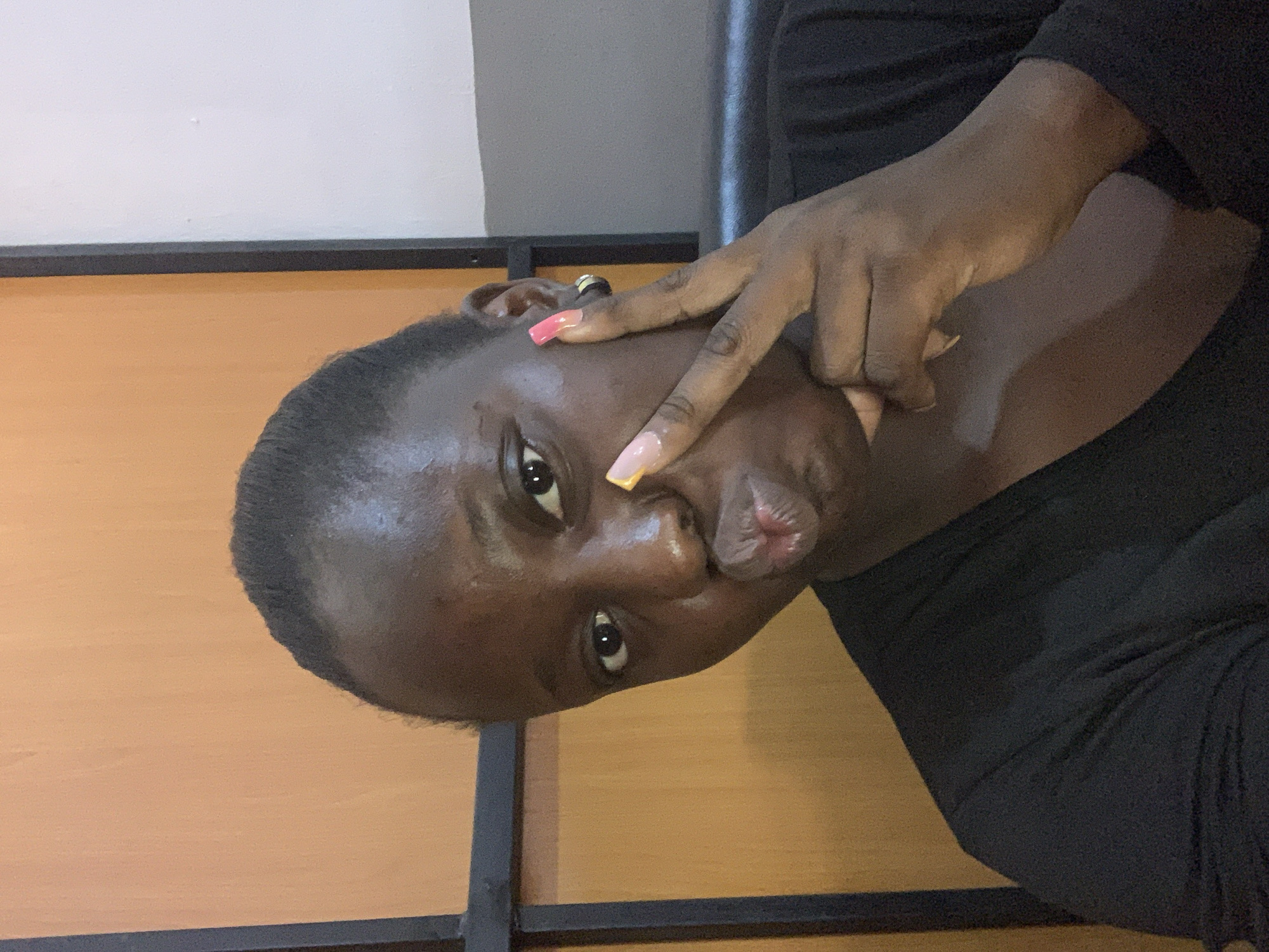 A young Black woman with dark skin and dark hair pulled back is pictured in a close-up, selfie-style shot. She is wearing a black top and has long, colorful acrylic nails. She is making a kissy face and has two fingers near her mouth, making a peace sign. The background appears to be a room with an orange wall and a black frame or structure. The lighting is warm and somewhat dim.