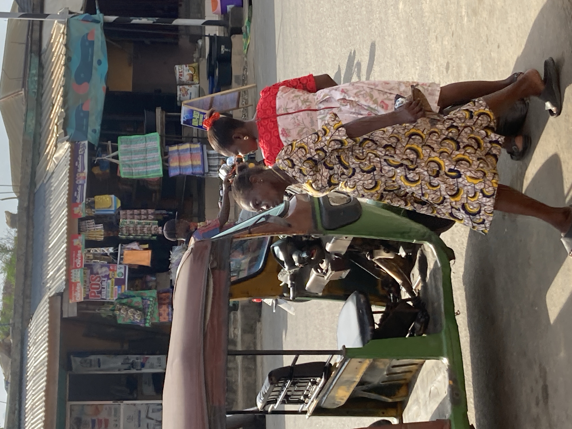 The image shows a street scene in Aba, Nigeria. Two women are seated on a green tricycle, angled toward the right of the frame. The woman closer to the viewer wears a yellow and black patterned dress; the other woman wears a white dress with red floral accents. The tricycle is parked next to a building; the building has a corrugated metal roof and an array of goods on display, including packaged foods, drinks, and advertising posters, some showing Chivita products. The street surface is paved and appears dry. A small boy stands in the background between the ladies.