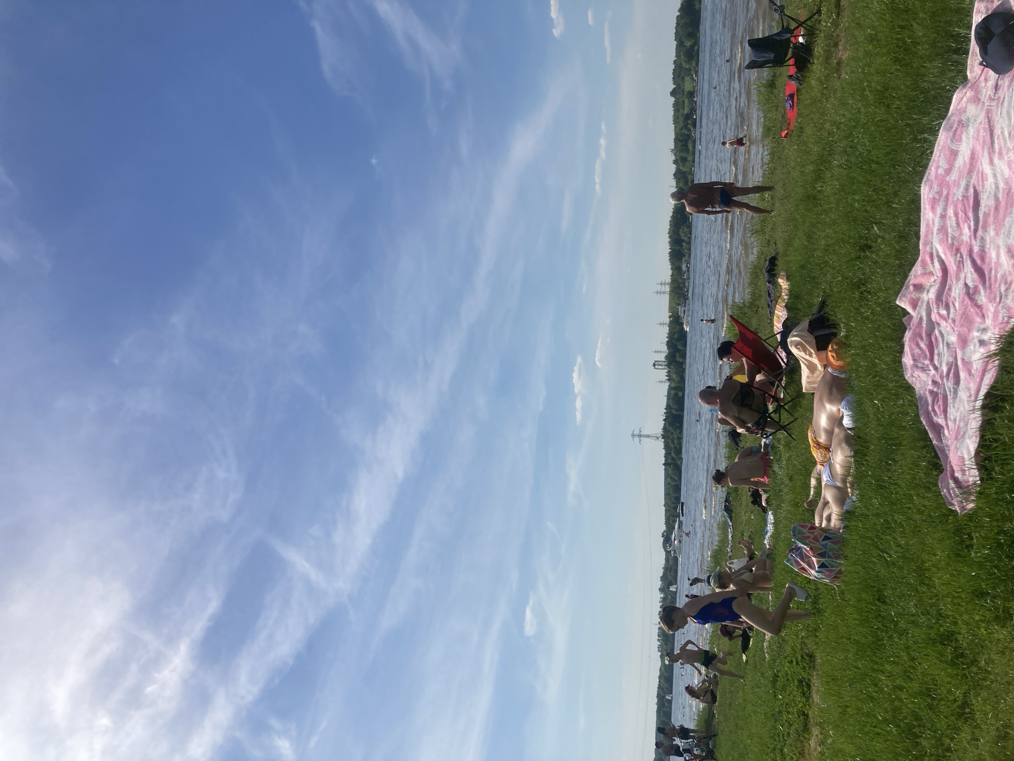 The image depicts a sunny summer day at a recreational area alongside a large body of water, identified as being in Kapustino, Russia. The scene features a grassy bank in the foreground sloping down to the water, where numerous individuals are engaged in leisure activities.
The sky is bright blue with scattered, wispy cirrus clouds, predominantly on the left side, indicating clear weather. The sun is high, suggesting midday or early afternoon.
On the grassy bank, approximately twenty people are visible. In the immediate foreground to the right, a pink and white patterned towel is spread on the grass. To its left, two individuals are lying prone, sunbathing; one wears yellow and white swimwear. A colorful geometric-patterned tote bag rests near them. Moving left, a young person in a blue one-piece swimsuit walks towards the water, while another young person in blue shorts and a light top walks further to the left. Other individuals are seated on the grass or on red and black folding chairs. To the far right, a bald, shirtless adult male in blue swim trunks stands at the water's edge, facing the water.
In the water, several people are wading or swimming. A small figure in red trunks is seen wading further out near the standing man. The water itself is rippled, suggesting a slight breeze or current.
Across the expanse of water, a distant treeline marks the opposing bank. Several boats are present on the water, including a large white yacht-like vessel in the mid-distance and smaller sailboats or motorboats further out. In the background, beyond the distant treeline, several power transmission towers and associated power lines are visible, indicating industrial or semi-urban infrastructure.