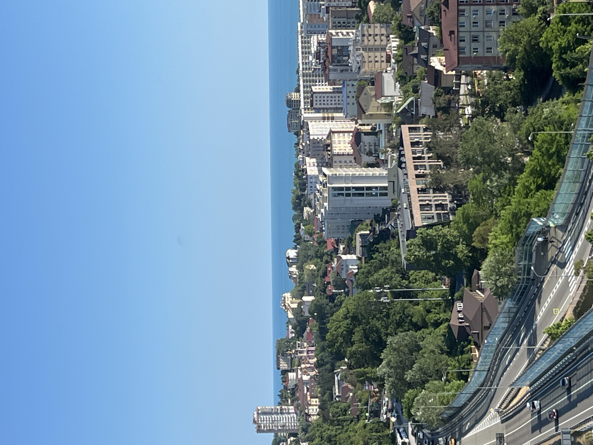 The image shows a panoramic view of a coastal city, likely Sochi, Russia, on a clear day. The foreground features a multi-lane highway with a few cars driving on it. A blue transparent fence runs along the side of the highway. Beyond the highway, the scene transitions to a densely populated urban area with a mix of residential buildings and taller apartment complexes, interspersed with lush green trees and vegetation. In the background, the calm blue sea stretches to the horizon, meeting a clear blue sky. The architecture of the buildings suggests a modern development, with several high-rise structures visible among the lower buildings. Some older buildings with distinctive architectural features, possibly religious or historical sites, are also present. The overall impression is of a vibrant city situated between a highway, a verdant landscape, and the sea.