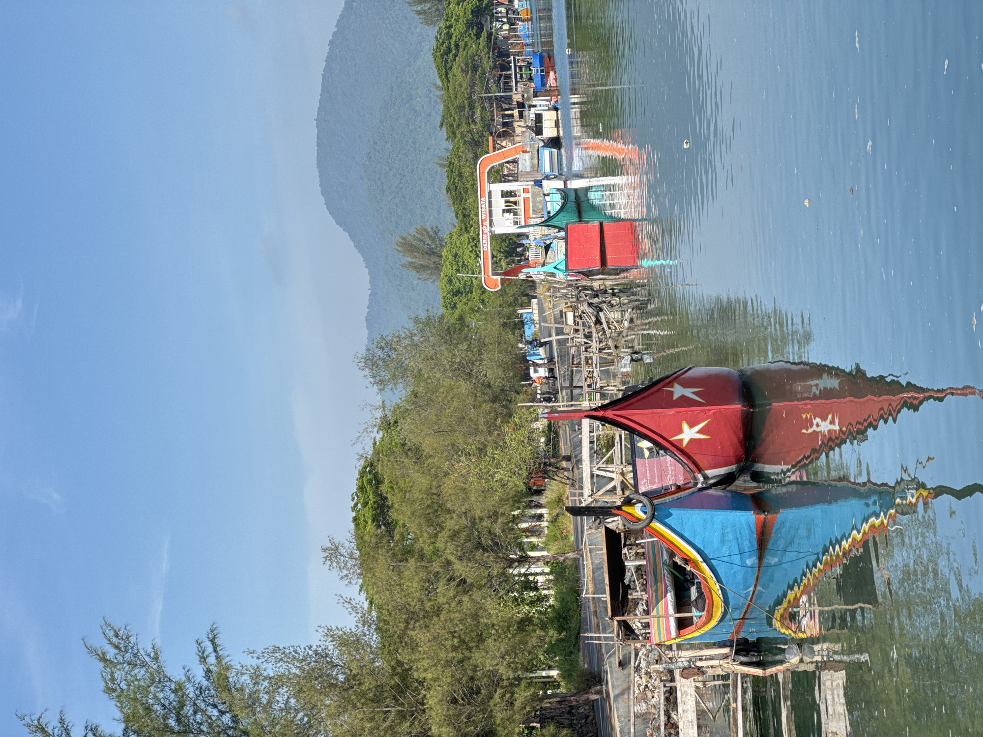 This is a medium-shot, landscape-oriented photograph taken from a slightly elevated perspective, looking across a calm body of water towards a waterfront. The scene depicts a small harbor or pier area, likely in Banda Aceh, Indonesia, given the Indonesian text visible on signage.
In the foreground, two vibrantly colored boats are moored. The boat on the left is primarily blue with yellow, red, and green accents along its upper edge. It has a distinct, pointed prow. The boat on the right is red with two white star-shaped emblems on its front. Both boats are reflected in the still water, creating a symmetrical mirror image. The water is a calm blue-grey, with small ripples and scattered debris floating on its surface.
Behind the foreground boats, a wooden pier or walkway is visible, constructed from weathered planks and pilings. Further back, a row of trees with dense green foliage lines the shore, creating a backdrop of natural greenery. Beyond the trees, a misty, blue-hued mountain range rises in the distance under a clear, pale blue sky.
On the waterfront, several other boats are moored, including a green and blue boat in the middle distance. A distinctive orange and white structure with Indonesian text that appears to read "DERMA WISATA" (Tourist Wharf) is also present, suggesting a tourist-oriented area. Small buildings and more wooden structures are scattered along the shoreline. In the background, a few people are visible, some near the boats and structures, and one person on a bicycle further down the road, indicating some human activity. The overall atmosphere is serene and tropical.