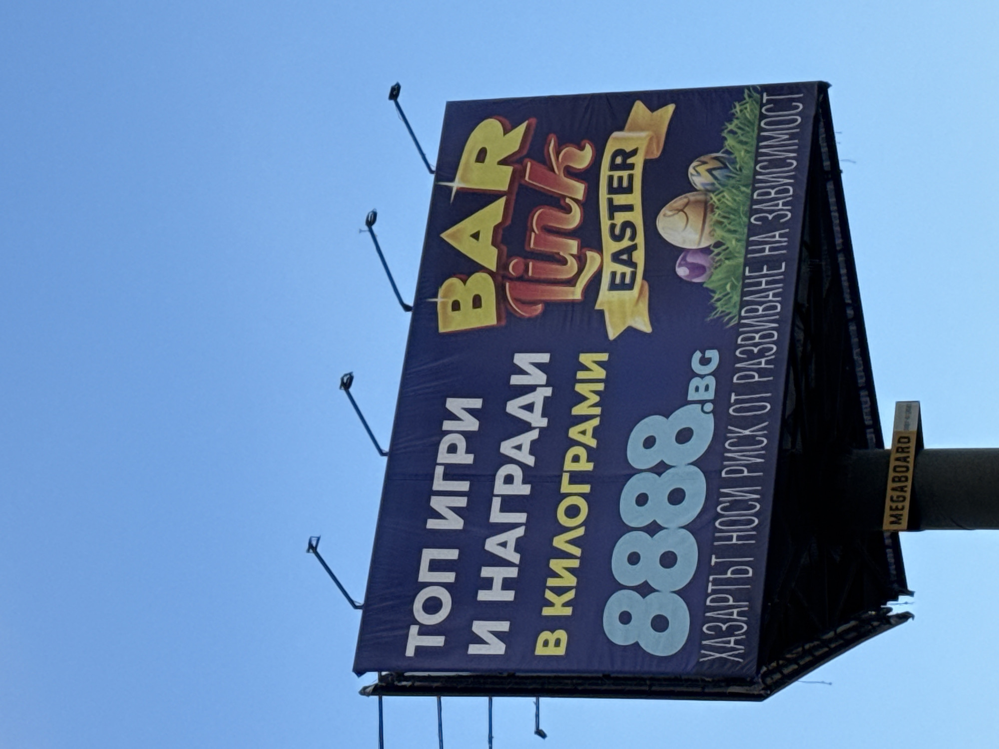 The image shows a billboard advertising "Bar Link" Easter promotions, with the text "TOP GAMES AND AWARDS IN KILOGRAMS" and the phone number "8888.BG". The text "GAMBLING CARRIES A RISK OF DEVELOPING DEPENDENCE" is also present. The billboard appears to be located outdoors in Sofia, Bulgaria, based on the Bulgarian text. The billboard is mounted on a metal frame against a clear blue sky. A "MEGABOARD" tag is visible on the side of the structure.