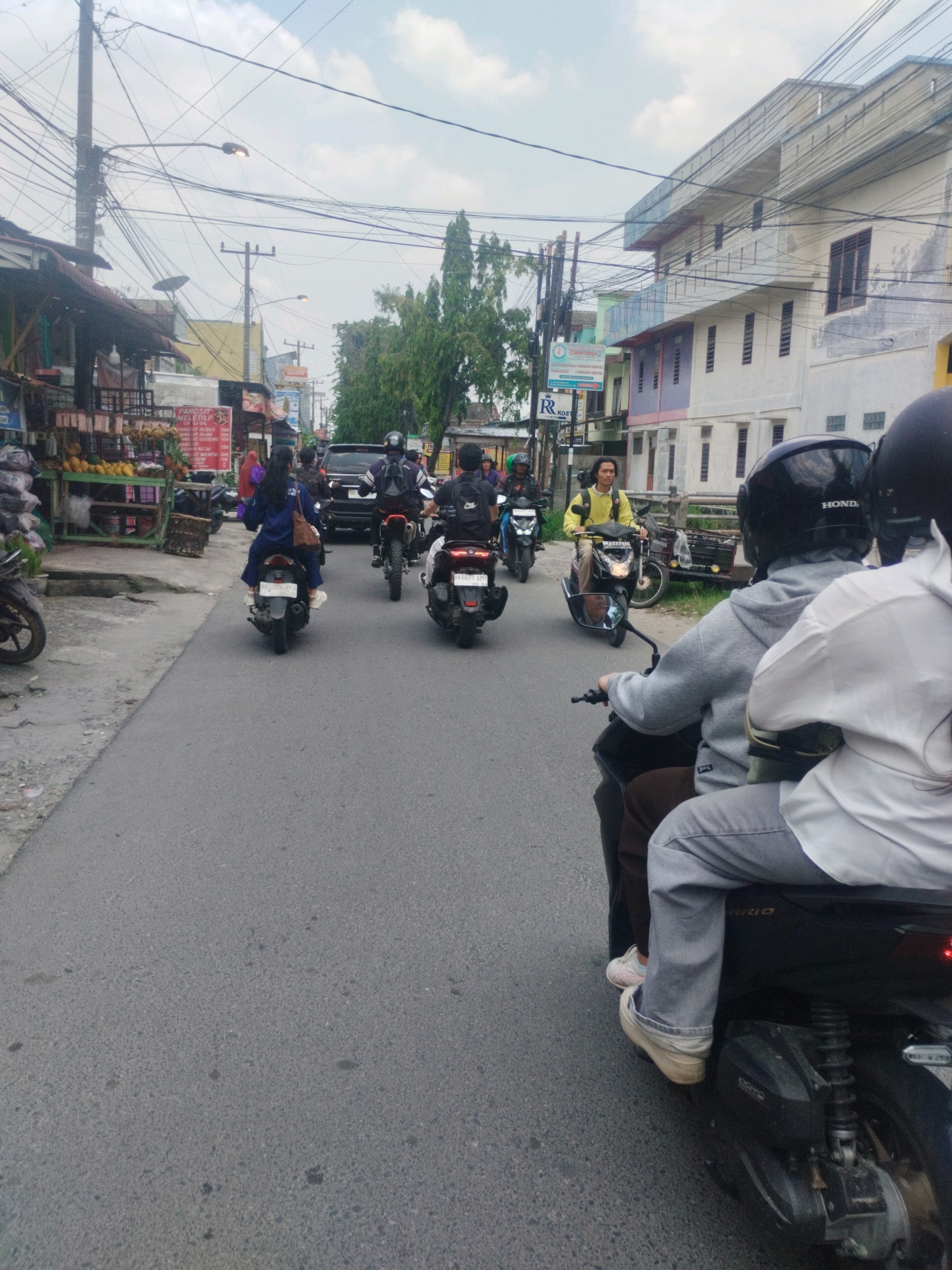 In City of Medan, Indonesia, a group of people are riding motorcycles down a street lined with shops and buildings. In the foreground, two people on a motorcycle are visible from behind. The person in front is wearing a grey hoodie and grey pants, and the person in the back is wearing a white shirt and grey pants. Both are wearing helmets. The motorcycle has the brand name "HONDA" on the helmet of the person in front. The street is paved and appears to be a typical urban road. On the left side of the street, there are several shops with merchandise displayed outside. On the right side of the street, there are multi-story buildings. Several other motorcycles are also visible on the road, some with multiple riders. The sky is overcast with some clouds. The overall scene depicts a busy street with common mode of transportation in this region.
