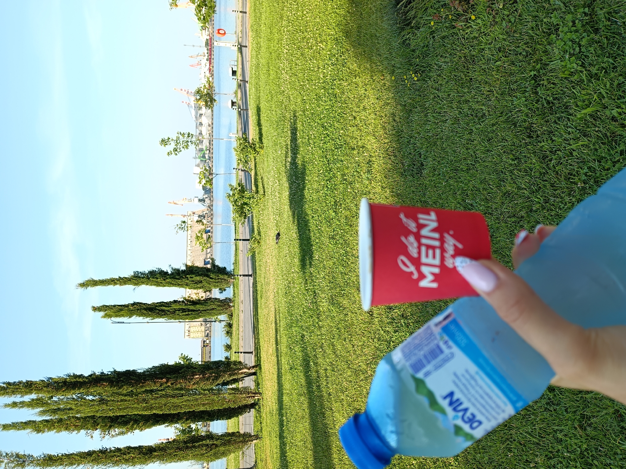 This is a close-up, point-of-view shot of a hand holding a blue plastic bottle of Devin mineral water and a red disposable cup. The bottle's label is partially visible, showing the brand name "DEVIN" and "500ml." The red cup has white text that reads, "I do it MEINL way." The background shows a green lawn, a body of water, and a city skyline with industrial cranes, typical of the Port of Burgas, Bulgaria. The sky is clear and blue. The lighting suggests it is daytime, with sunlight casting shadows on the grass.