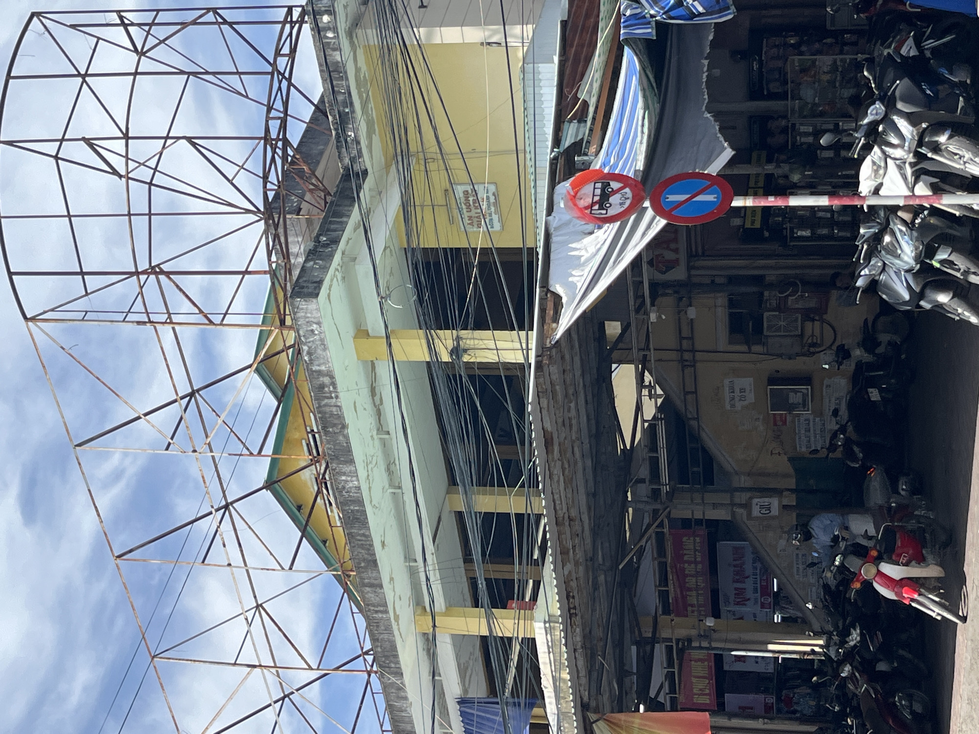 A street scene in Nha Trang Ward, Vietnam, depicts a building with multiple levels and a large, rusted metal framework for a billboard on its rooftop. The building's facade is painted yellow, with exposed concrete on the upper edges. Power lines are strung haphazardly across the front of the building at various levels. Below the upper levels, a covered walkway or market area is visible, supported by metal beams and covered with corrugated metal and fabric tarps. Parked motorcycles are lined up in the foreground. A person wearing a light blue shirt and a helmet is sitting on a red and white motorcycle. Two traffic signs are mounted on a pole: a circular red and white sign with a bus symbol and text, and a blue and white circular sign with a diagonal red bar. In the background, several shops are visible, some with signs in Vietnamese. The sky is partly cloudy.