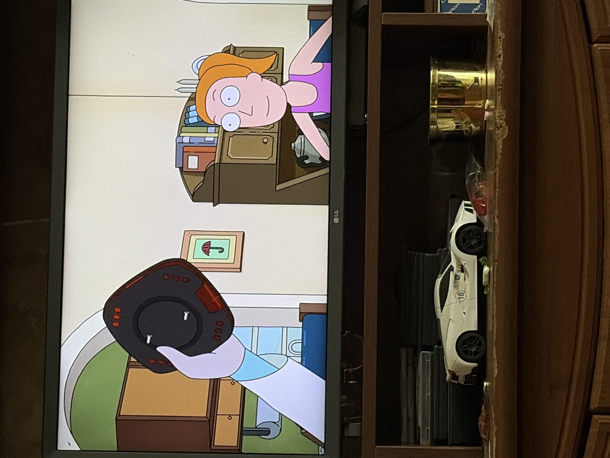 A television screen displays an animated scene from Rick and Morty. A white-gloved hand, belonging to Rick Sanchez, holds a black, square-shaped device with red lights and buttons. Rick is wearing a blue and white lab coat. In the background, Summer Smith, with orange hair and a purple tank top, sits at a desk with books. To the left of Summer is a framed picture of an umbrella. The television is placed on a wooden shelf unit. On the shelf below, a white toy sports car with the number 18 on its side is visible, along with a gold-colored container. The scene is indoors, in an unspecified location.