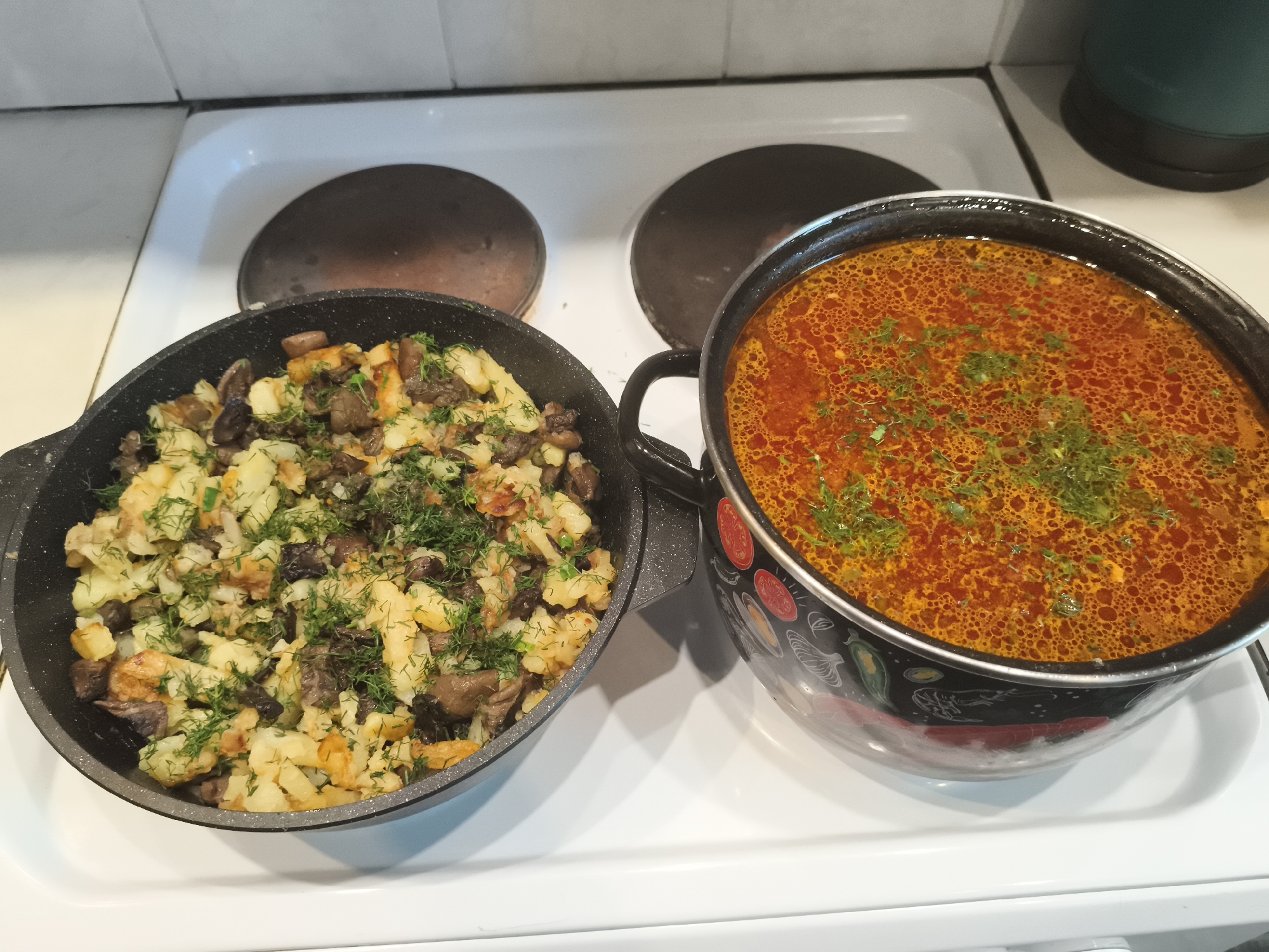An overhead shot shows two cooking pots on a white electric stovetop. The pot on the left is a black frying pan filled with fried potatoes and mushrooms, garnished with chopped dill. The pot on the right is a larger black pot with a colorful design, containing a vibrant orange soup, also garnished with dill. The stovetop has two dark electric burners. The background shows a white tiled wall. The scene is consistent with a domestic kitchen.