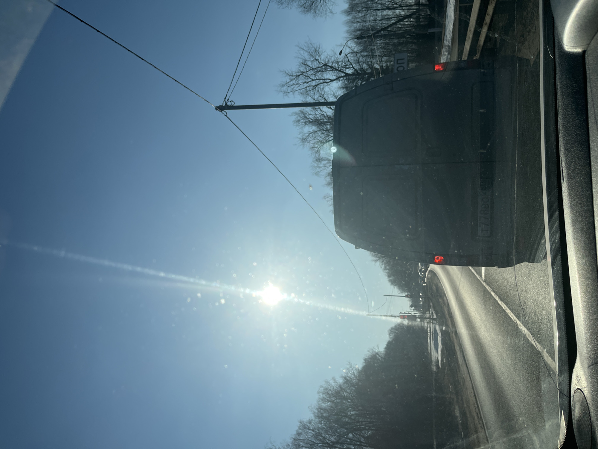 The image depicts a daytime view from the interior of a vehicle traveling on a multi-lane asphalt road in Rogachovo, Russia. A clear, bright blue sky with a prominently visible sun in the upper-left quadrant generates significant lens flare and glare on the observer's windshield, which also shows multiple bright spots consistent with dust or debris. Directly ahead, a dark-colored commercial van, possibly grey or black, occupies the same lane. Its rear is visible, including an illuminated red taillight on the right side and a partially obscured Russian license plate beginning with "T778BO." The road surface appears reflective under the sunlight, with visible white lane markings. Bare deciduous trees line both sides of the road, and patches of snow are visible on the shoulders and adjacent ground. Utility poles with overhead lines are present along the right side of the road, where a white sign with Cyrillic text, including "ОП," is partially visible behind the van. Other vehicles are faintly discernible in the distance down the road. The top of the observer's vehicle dashboard is visible in the lower foreground. No people are explicitly visible.