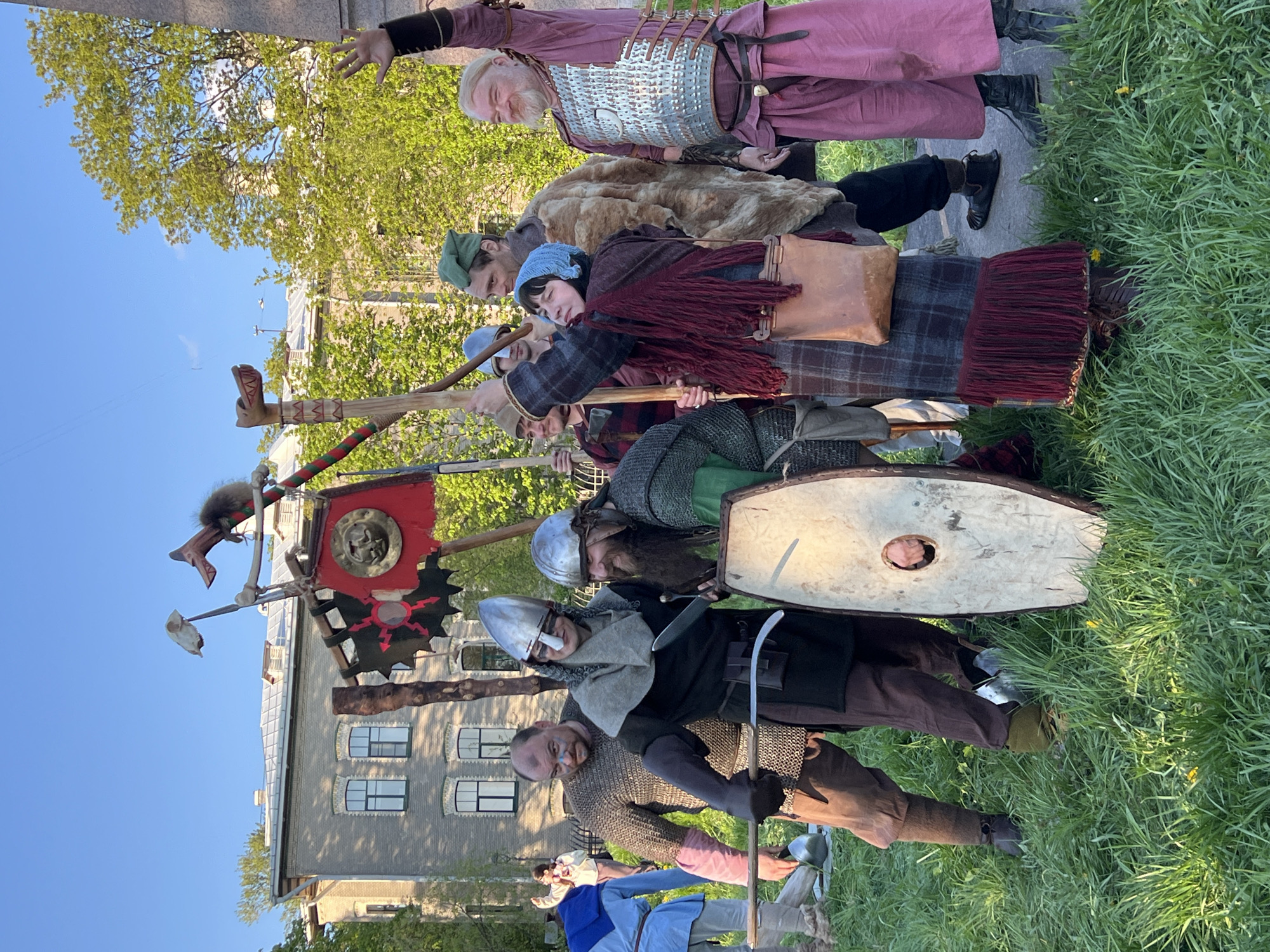 A group of seven people dressed in historical clothing, resembling medieval attire, are standing in a line outdoors on a grassy area. They are positioned in front of a light-colored building with multiple windows and under a partially visible blue sky. One person at the front of the line wears a pink robe and silver armor-like pieces on the chest. Another person wears a fur coat. Other individuals are dressed in various shades of browns and greys with helmets and chainmail, some holding spears, shields, and a banner. The banner has red and black design elements and appears to be attached to a pole decorated with animal parts. The scene takes place in Saint Petersburg, Russia.
