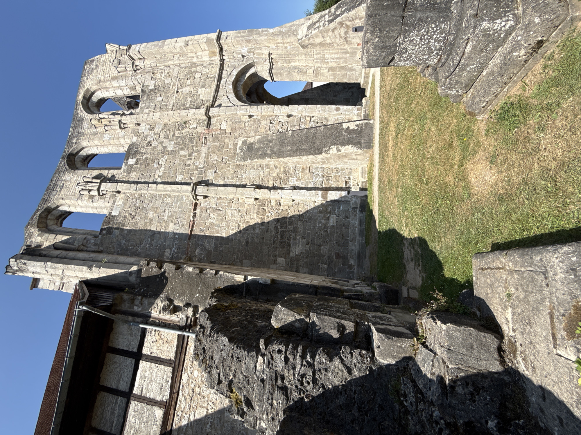 The image shows the ruins of Walkenried Abbey, a former Cistercian monastery located in Walkenried, Germany. The ruins are constructed of large, weathered stone blocks and feature Gothic architectural elements such as arched windows and buttresses. A portion of a building with a brown tiled roof and timber framing is visible to the left, attached to the stone ruins. The foreground is dominated by large, rough-hewn stone blocks, some of which appear to be fallen architectural elements. Green grass and dry, sun-baked ground cover the area around the ruins. The sky is a clear, bright blue, indicating a sunny day. There are no people or animals visible in the image. The scene is static, depicting the enduring stonework of the historical site under a clear sky.