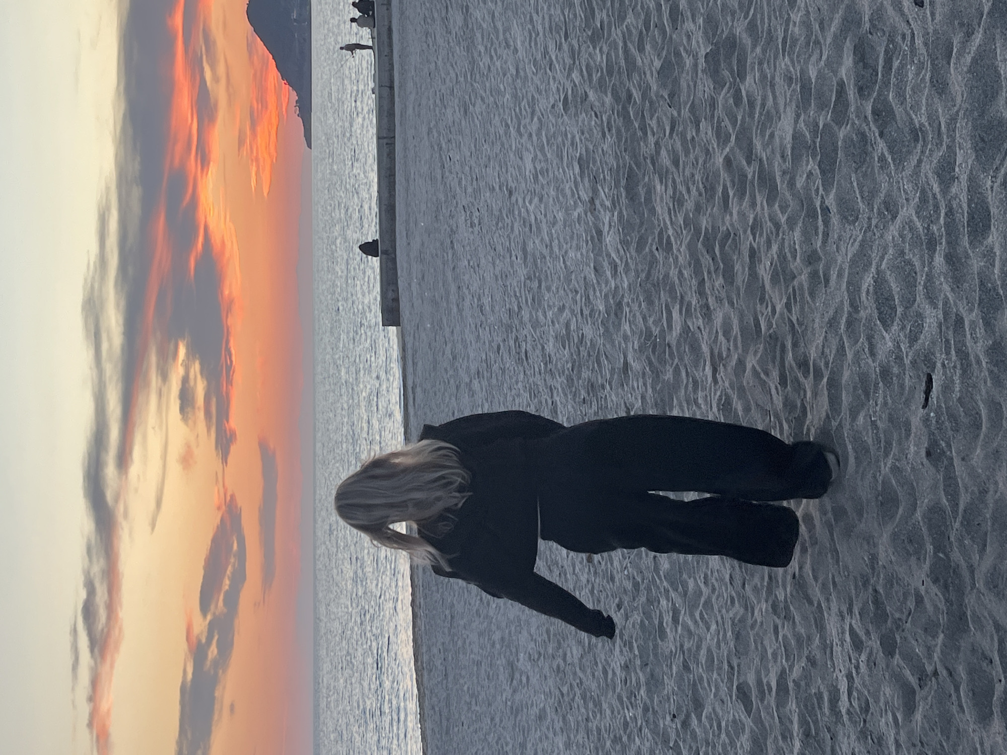 The image captures a serene coastal scene at sunset, likely in Alanya, Turkey. In the foreground, a person with long, blonde hair, dressed in dark attire, stands on a sandy beach, facing away from the camera. They are looking out towards the sea and the horizon, with their back to the viewer. The sand beneath their feet features distinct ripple patterns, suggesting the movement of water or wind.
To the left, the calm, shimmering surface of the sea extends into the distance. A concrete pier or structure runs horizontally across the midground, separating the beach from the open water. Several small, silhouetted figures, likely other people, are visible on this pier in the background, some possibly engaged in fishing or observing the view.
The sky is a prominent feature, showcasing the dramatic colors of a sunset. Vivid streaks and patches of orange, red, and yellow clouds are interspersed with cooler grey and purple hues, creating a striking and colorful backdrop. A dark, silhouetted mountain range or landmass is visible on the far left, bordering the brightly colored sky. The overall impression is one of tranquility and natural beauty, with the warm light of the setting sun illuminating the clouds. No text is visible in the image.