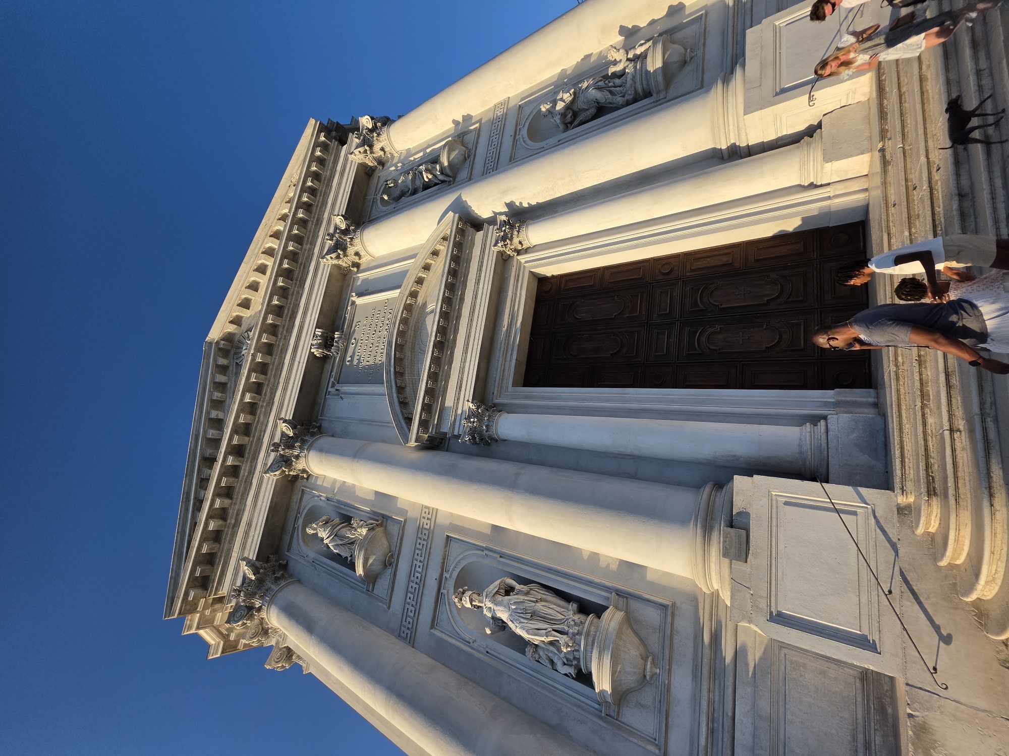 The image captures the facade of the Church of San Giorgio Maggiore in Venice, Italy, viewed from a low angle against a clear blue sky. The imposing neoclassical architecture is characterized by large white columns, intricate sculptures in niches, and a grand wooden entrance. Sunlight casts long shadows across the building and the surrounding stairs. Several individuals are visible on the steps leading up to the church, some walking and one man talking on his phone. A dog is also present on the stairs. The scene appears to be a tourist attraction, with people observing and interacting with the historic structure.