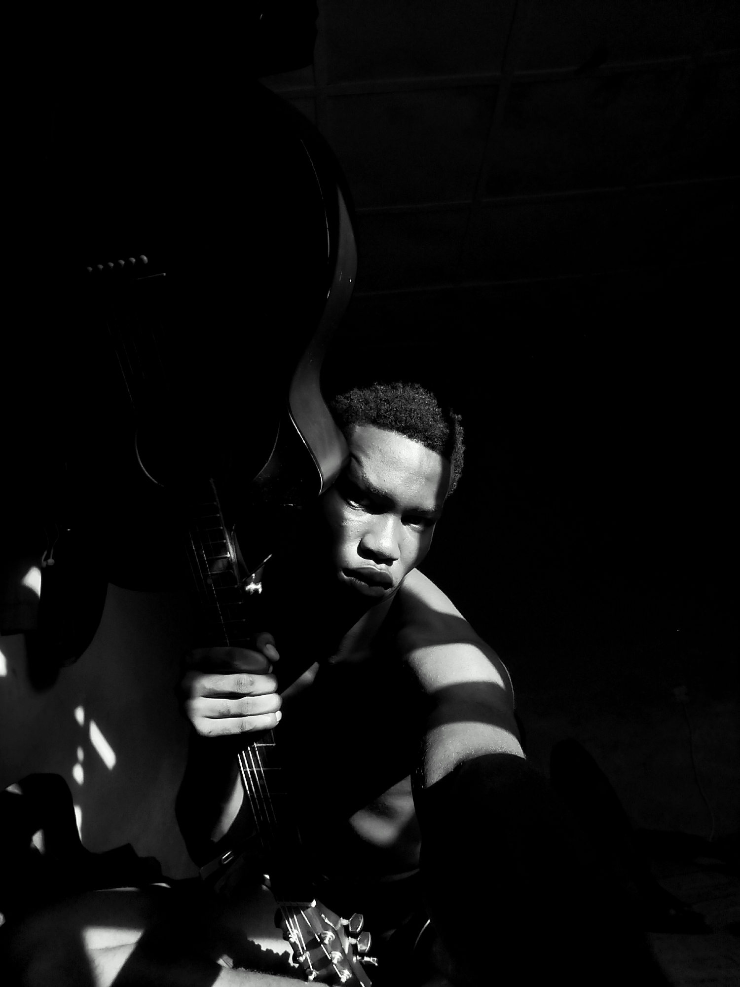 A high-contrast, black and white portrait features a young man holding an acoustic guitar in a dimly lit indoor setting in Keffi, Nigeria. The man, likely in his late teens or early twenties, has short, dark, curly hair and appears shirtless or wearing dark clothing. He is illuminated by a strong, directional light source that creates dramatic chiaroscuro effects, highlighting parts of his face—including his forehead, nose, and cheekbone—and his right arm and shoulder. His expression is serious and contemplative, with his gaze directed slightly downwards.
He cradles a dark acoustic guitar upright, with his right hand gripping the neck near the fretboard. The upper bout and headstock of the guitar are visible, largely silhouetted against the dark background, with some details of the bridge pins and tuning pegs discernible. Diagonal patterns of light and shadow fall across his arm and body, suggesting sunlight streaming through a window or blinds.
The background is predominantly dark, almost entirely black, emphasizing the stark contrast between light and shadow. The moody lighting suggests either early morning or late afternoon. No discernible text is visible in the image.