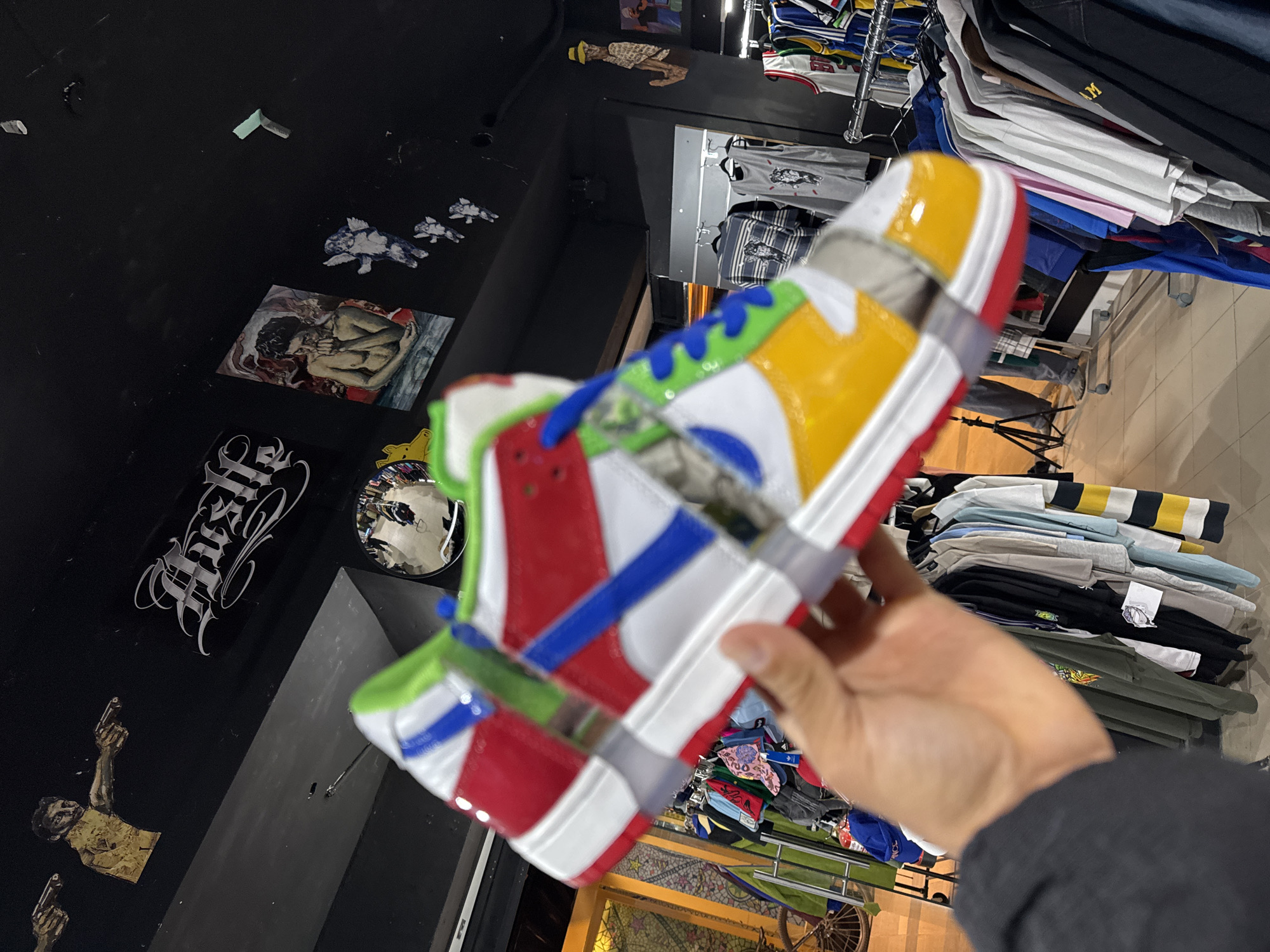 A hand holds a colorful sneaker in a store in Barcelona, Spain. The shoe is brightly colored with red, white, blue, green, and yellow sections and is held together with gray bands. The background includes a wall with various stickers and a small mirror. To the right of the sneaker, there are racks of clothing. The lower part of the frame shows flooring with a mosaic pattern.