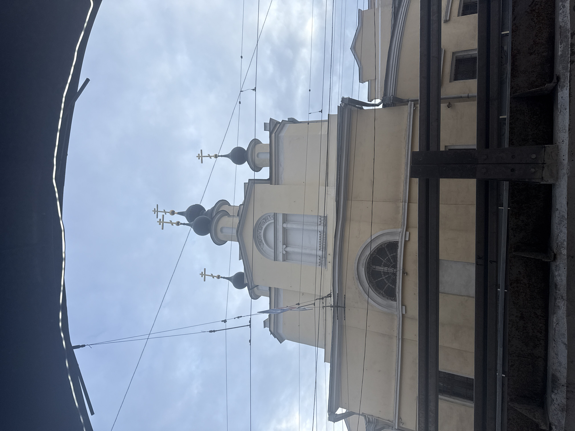 The image shows a church with multiple domes and crosses, likely an Orthodox church, against a sky with overcast conditions. The church is built in a traditional style with rounded domes and arched windows. Power lines are visible in the foreground. Metal construction or support beams are visible in the bottom right corner of the image. The image is likely taken from a lower vantage point looking upwards. Based on the architecture and overall environment, the image is likely of the Church of St. Nicholas in Pyzhy, Moscow, Russia.