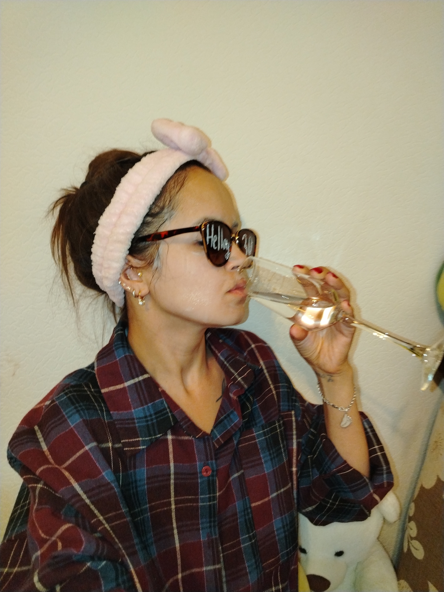 A woman is seen from the chest up, wearing a pink fluffy headband with a bow, sunglasses with "Hello 30" written on them, and a red, blue, and white plaid shirt. She is holding a champagne flute filled with a clear liquid and drinking from it. She has multiple earrings in her left ear, a bracelet on her right wrist, and a tattoo on her left forearm. In the background, a white stuffed animal with black eyes and a brown nose is visible, along with a wall and a draped fabric. The location is not determinable from the image.