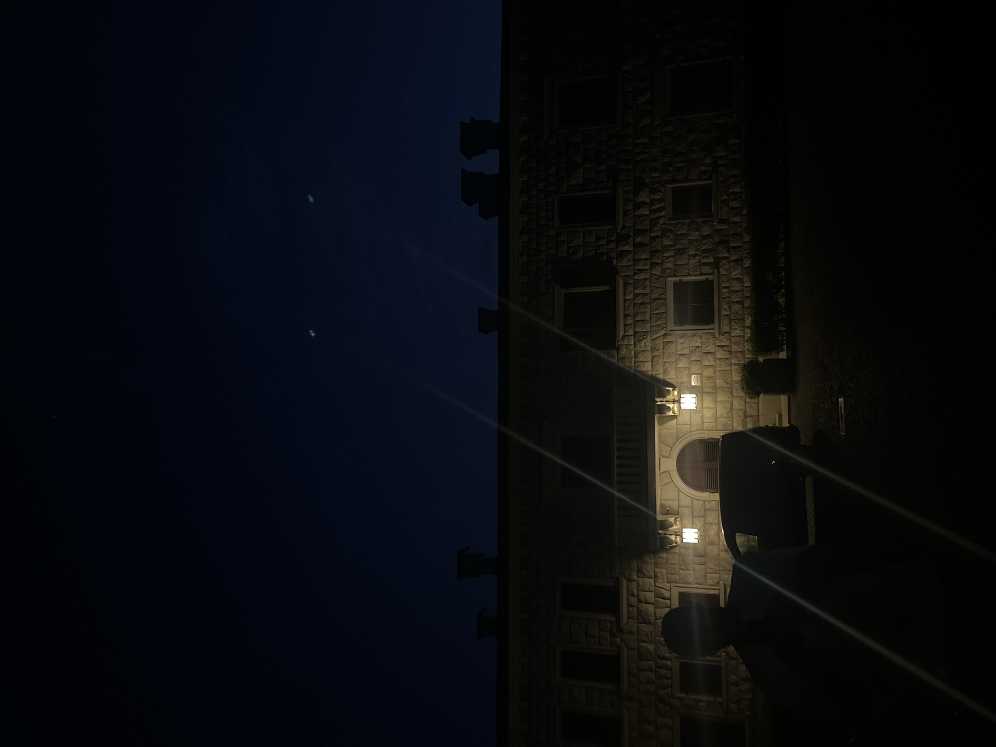 A nighttime shot depicts a stone building with lit windows and exterior lamps, set against a dark sky. The building's facade features a prominent arched doorway. A silhouette of a person and a vehicle are visible in the foreground. The location is in the city of Unknown, Unknown.
