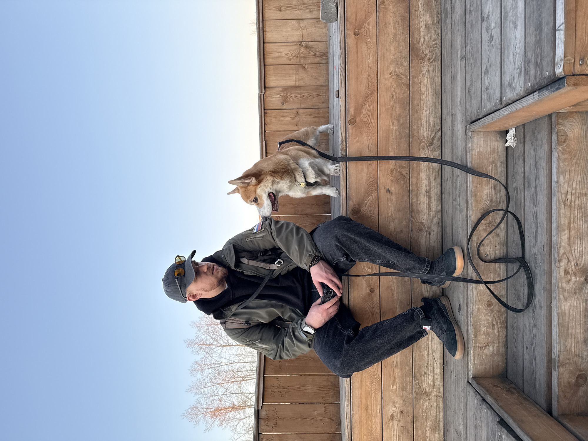 The image shows a man sitting on wooden stairs in Borzya, Russia, with a corgi standing on a raised wooden platform. The man is wearing a dark jacket, black jeans, and sneakers with a brown sole. He has a cap on his head and sunglasses perched on top of it. A black leash connects him to the corgi. The background includes a light blue sky and bare branches of a tree. The wooden structure appears to be a deck or patio area. Some graffiti is visible on one of the wooden panels.