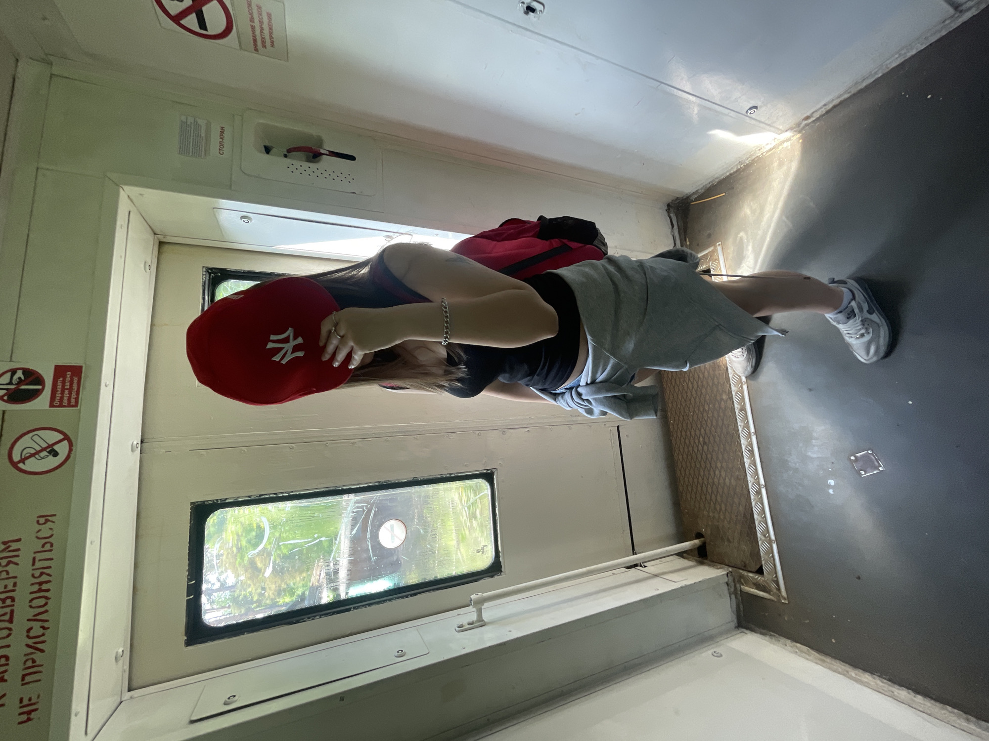 The image depicts the interior of a train vestibule, identified as being in Priozersk, Russia, by the Russian text on the signs. A single individual, appearing female, stands slightly off-center, facing away from the camera with her head turned to the left. She is obscuring her face with a red baseball cap featuring a white "NY" logo.
The person wears a black tank top, a grey sweatshirt tied around her waist, and grey and white athletic shoes. A red backpack is slung over her right shoulder. Her left hand, adorned with multiple rings, holds the cap. A silver chain bracelet is visible on her left wrist.
The train interior features light-colored walls and ceiling. A rectangular window on the left side shows a blurry view of green trees and foliage, suggesting the train is moving through a natural landscape. The window glass is visibly smudged. The floor is dark grey, with a section of metallic diamond plate flooring near a partially open train door on the left.
Multiple informational signs are affixed to the walls:
- At the top left, red Cyrillic text on a white background reads "К АВТОДВЕРЯМ НЕ ПРИСЛОНЯТЬСЯ" (DO NOT LEAN ON THE AUTOMATIC DOORS).
- Near the top center, two red-crossed-out circular symbols are visible. One depicts a cigarette (NO SMOKING), and the other shows a hand turning a handle, with accompanying text "Открывать двери вагона запрещено!" (Opening the car doors is forbidden!).
- On the right wall, a red emergency handle labeled "СТОП-КРАН" (STOP-CRANE) is positioned above a warning sign.
- Below the emergency brake, a partial sign reads "ВНИМАНИЕ ВЫСОКОЕ ЭЛЕКТРИЧЕСКОЕ НАПРЯЖЕНИЕ" (ATTENTION! HIGH ELECTRICAL VOLTAGE).
Natural light enters primarily from the left window, creating visible shadows on the floor and the lower right wall.