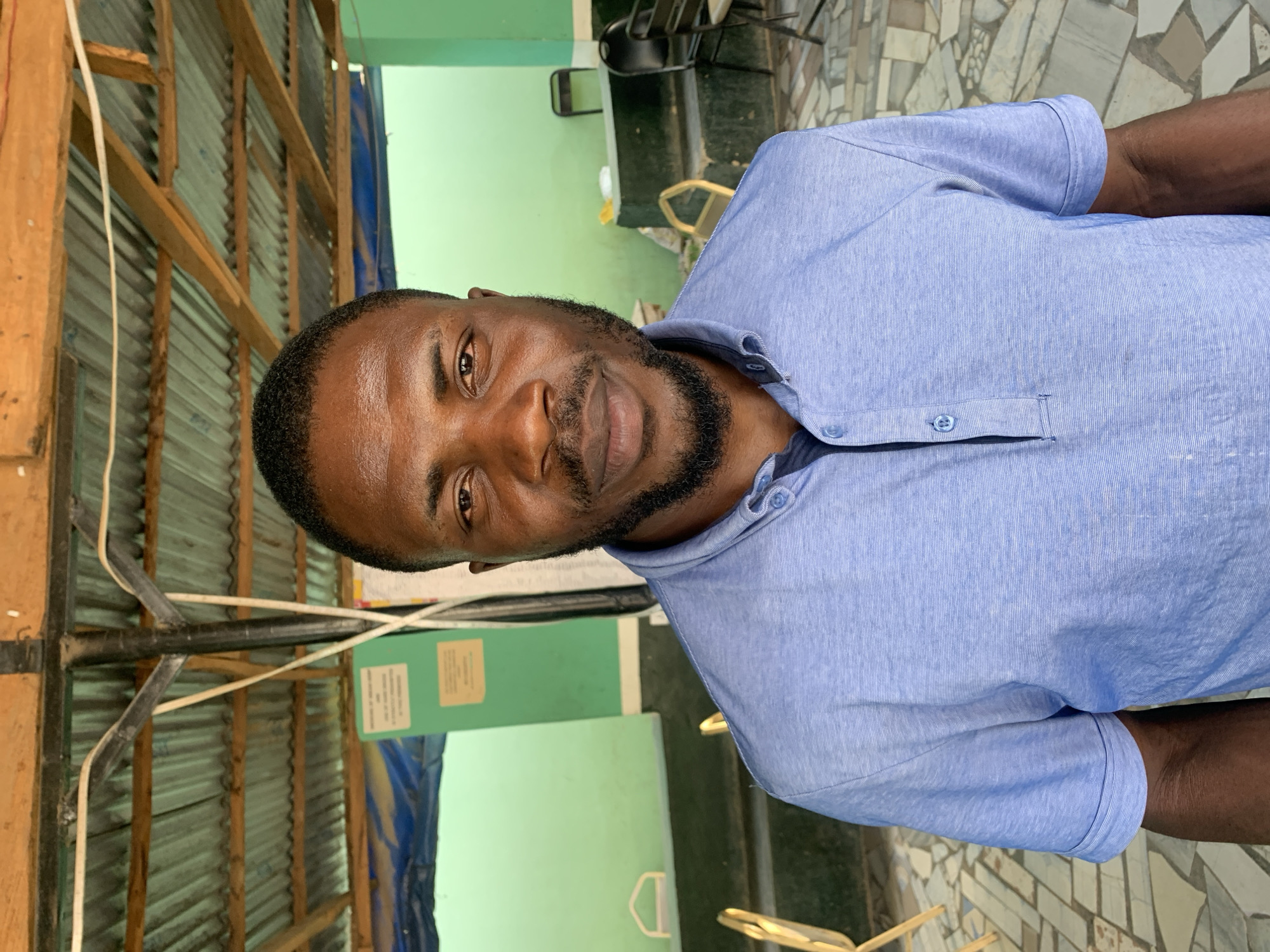 The image shows a man in a light blue button-down shirt. He has dark skin and a trimmed beard. The background reveals an environment with corrugated metal walls and wooden framing. A green painted wall section is also visible. Outdoor mosaic tile flooring suggests the location may be an outdoor or semi-outdoor space. The location is New Stadium, Nigeria. There are no apparent actions or interactions occurring in the photo.