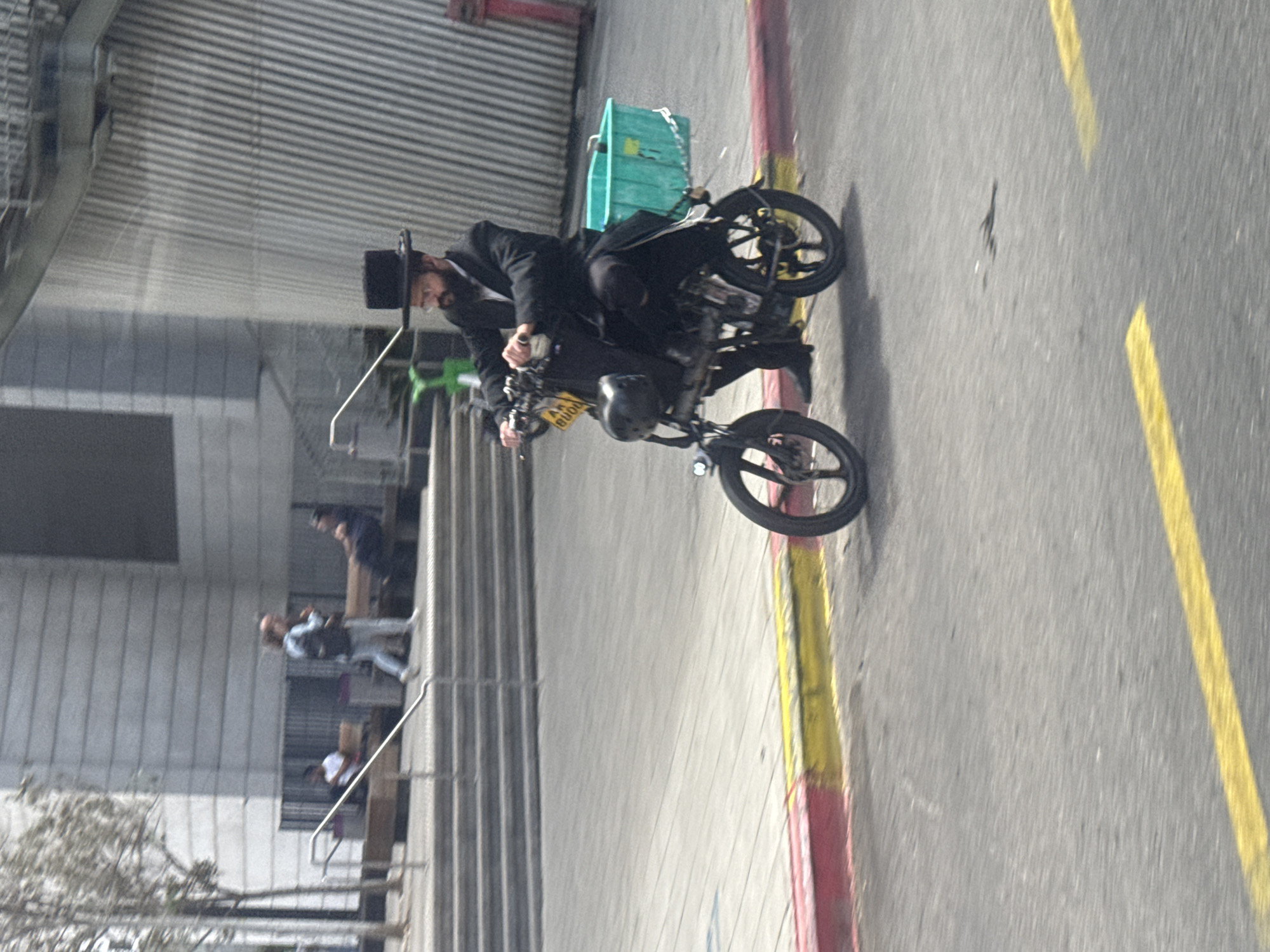 A man dressed in traditional Hasidic Jewish attire, including a black top hat and suit, rides a small, modern motorcycle on a city street. The motorcycle has a green basket attached. In the background, there is a building with horizontal siding, and several people are standing on a balcony. The road has yellow lines, with a red and yellow curb. The location is likely a neighborhood in Tel Aviv, Israel known for its Hasidic Jewish population.