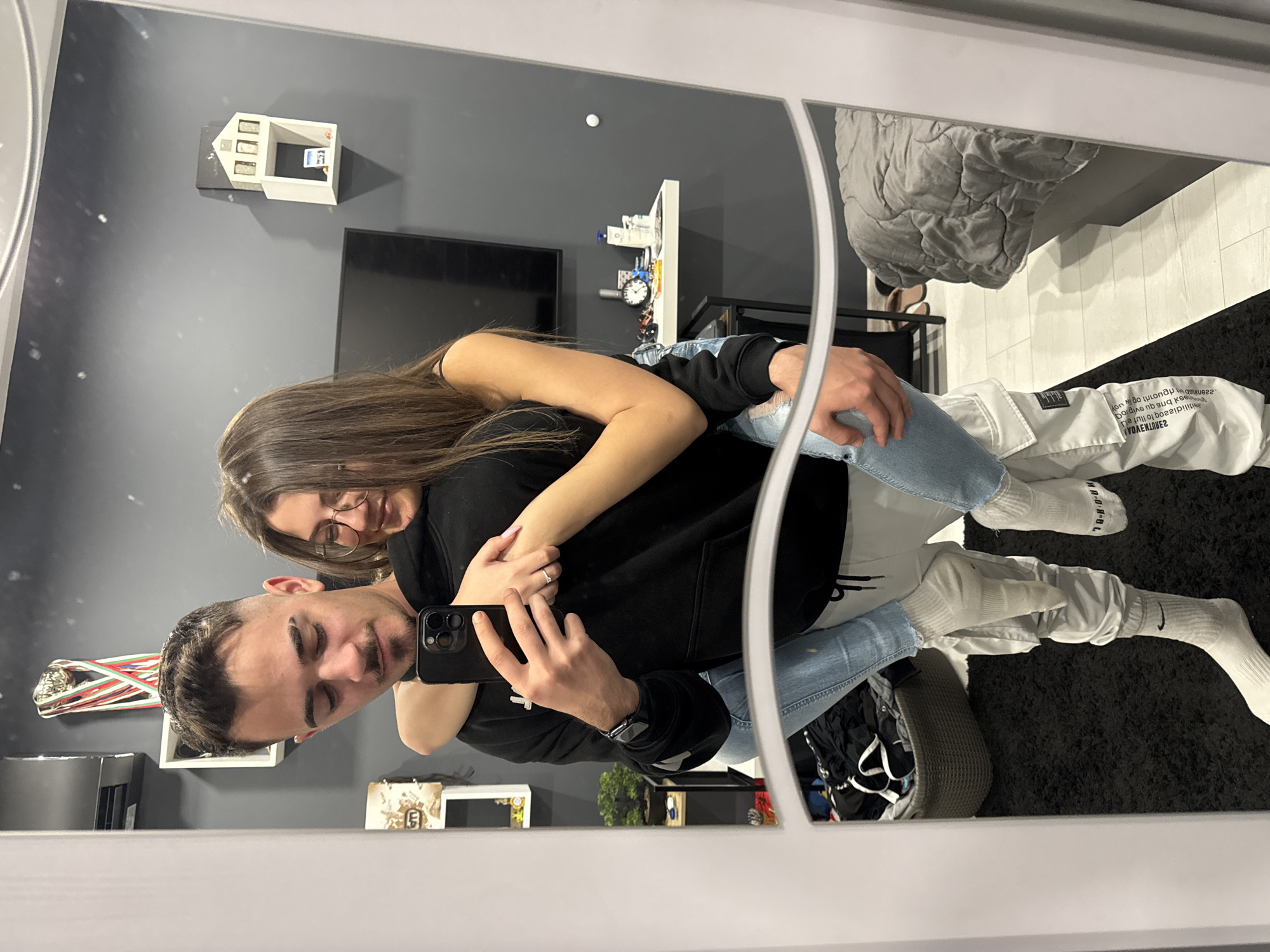 A couple takes a selfie in front of a mirror. The man is giving the woman a piggyback ride. The woman is wearing glasses and a black top. The man is wearing a black hoodie, ripped jeans and white pants over white socks. Both are wearing socks with the Nike logo. The man is holding a black smartphone. The setting appears to be a modernly decorated room with a gray wall. In the background, there's a television, shelves with various items, and a gray quilted blanket. It is unclear if this image was taken in Sofia, Bulgaria.