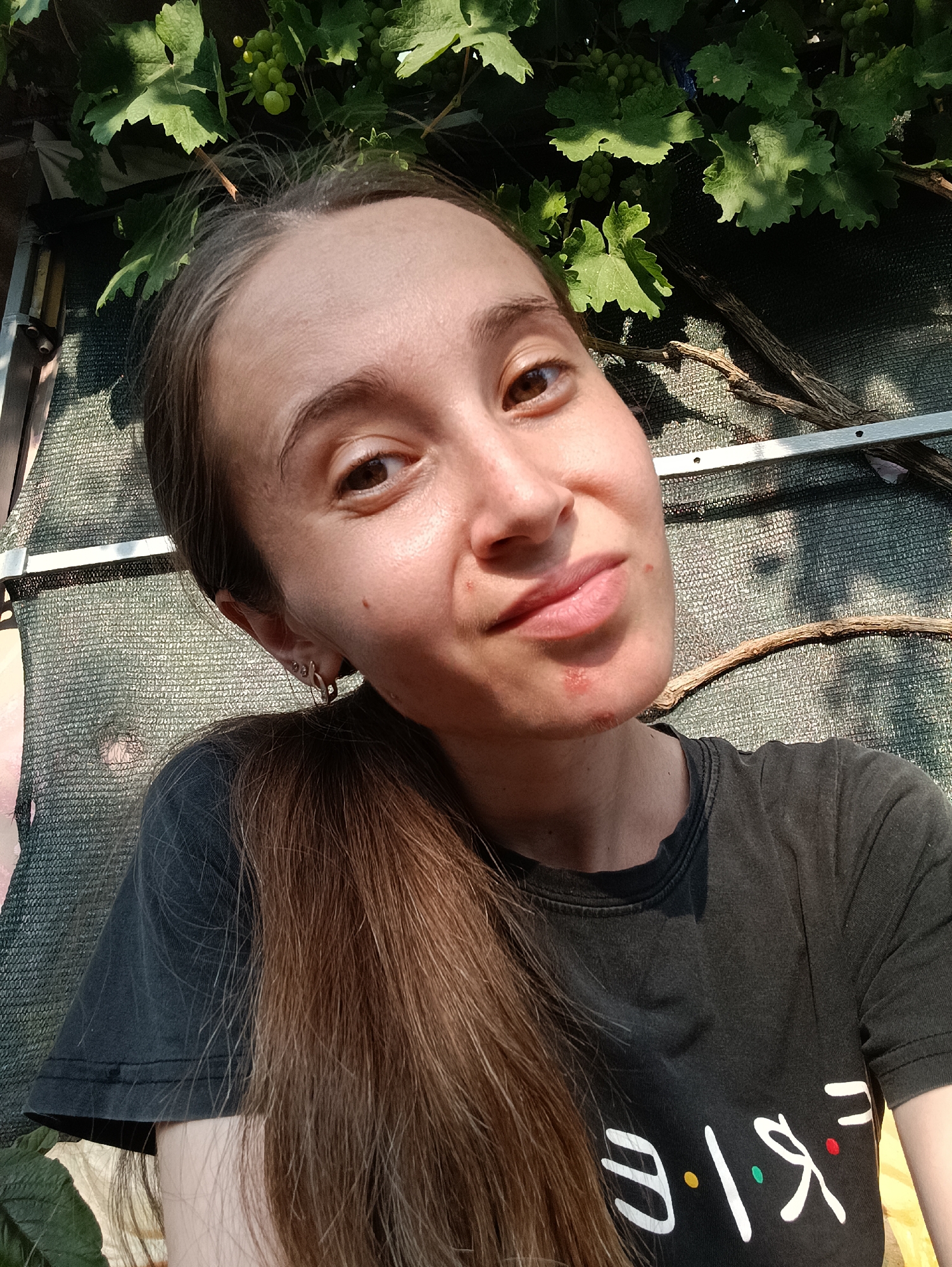 In a high-angle, close-up selfie, a young woman with long brown hair pulled to the side poses outdoors, with grapevines and leaves overhead and a green mesh fence in the background. The woman is wearing a dark t-shirt with a white logo that appears to say "Friends" with colored dots representing the letters. Her skin is slightly flushed, and she has a small red mark on her chin. The lighting suggests it is daytime, with sunlight filtering through the leaves and creating shadows. The setting, based on the provided location of Vypasne, Ukraine, is likely a rural or suburban area with a garden or vineyard.
