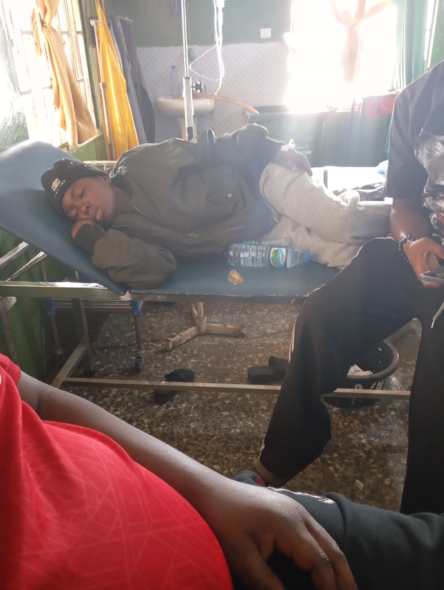 The image captures an indoor scene within what appears to be a medical facility in Jos, Nigeria. In the center, a young individual is lying on a blue medical examination table or stretcher, seemingly asleep or resting. They are dressed in a dark green jacket, light grey sweatpants, and a black beanie with "CELINE" written on it. An intravenous (IV) drip is connected to their left arm, with the clear fluid bag and stand visible behind them. A plastic water bottle, labeled "Swan," rests on the stretcher beside them.
Bright sunlight streams into the room from a window on the left, partially obscured by light-colored curtains, indicating it is daytime. In the background, a white sink or basin is mounted on the wall, adding to the medical setting cues. The floor is dark and textured.
Two other individuals are partially visible: one person wearing a black t-shirt and black pants is seated on the right, holding a mobile phone and wearing a beaded bracelet. Another person in a red t-shirt is visible in the bottom left corner of the frame. The overall atmosphere suggests a moment of rest or recovery during medical treatment.