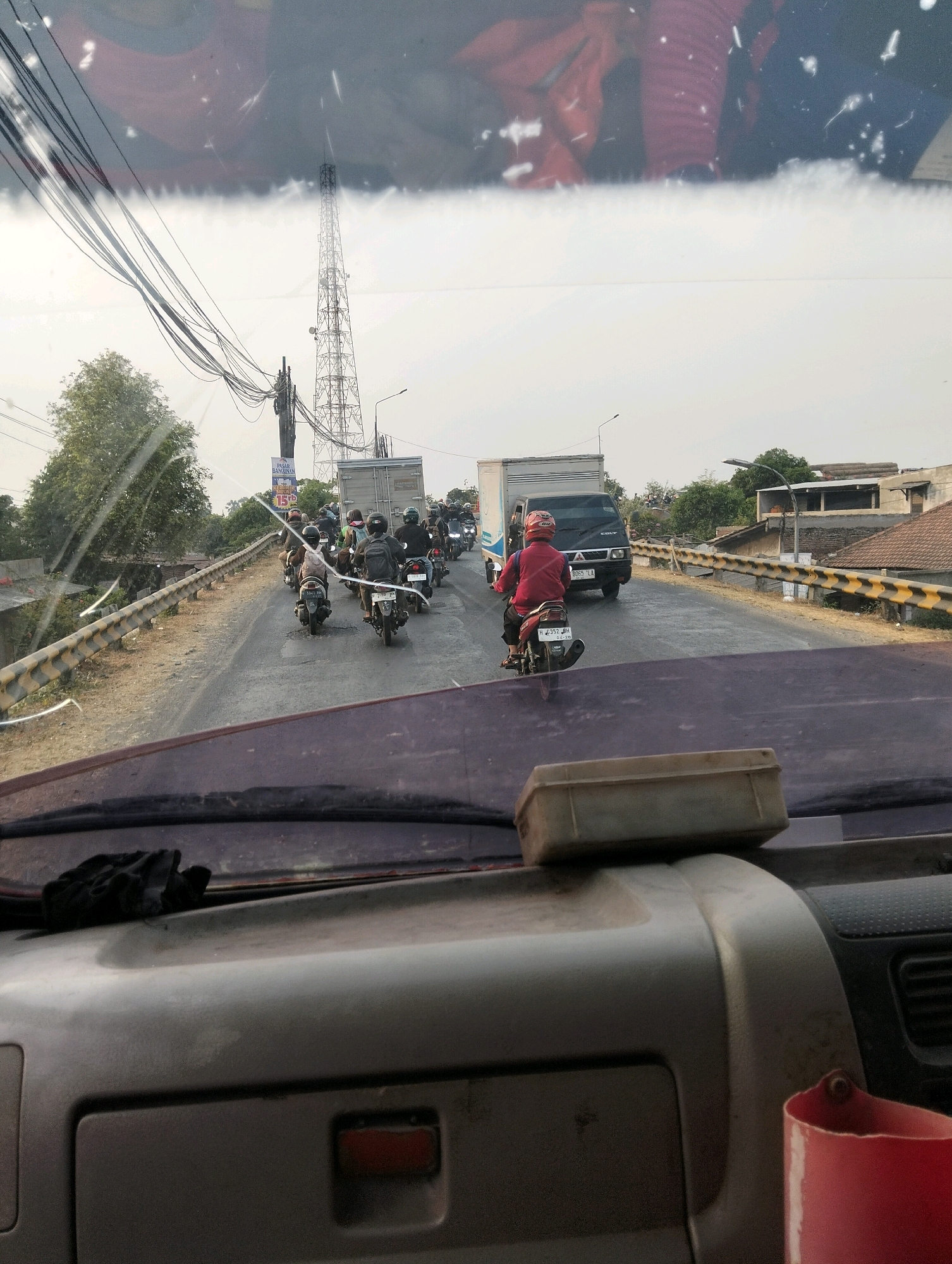 The image, taken from inside a vehicle looking forward, shows a traffic jam on a two-lane road, likely in Semarang, Indonesia. The road is lined with a yellow and black striped guardrail on the right side. In the distance, a tall communications tower is visible, along with a smaller tower structure. Several motorcycles are in the traffic jam, with riders wearing helmets. One motorcycle in the foreground has a rider in a red shirt and helmet. Two large trucks are also present in the traffic. The sky is overcast. The dashboard of the vehicle is visible in the foreground, with a dust-covered object placed on it.