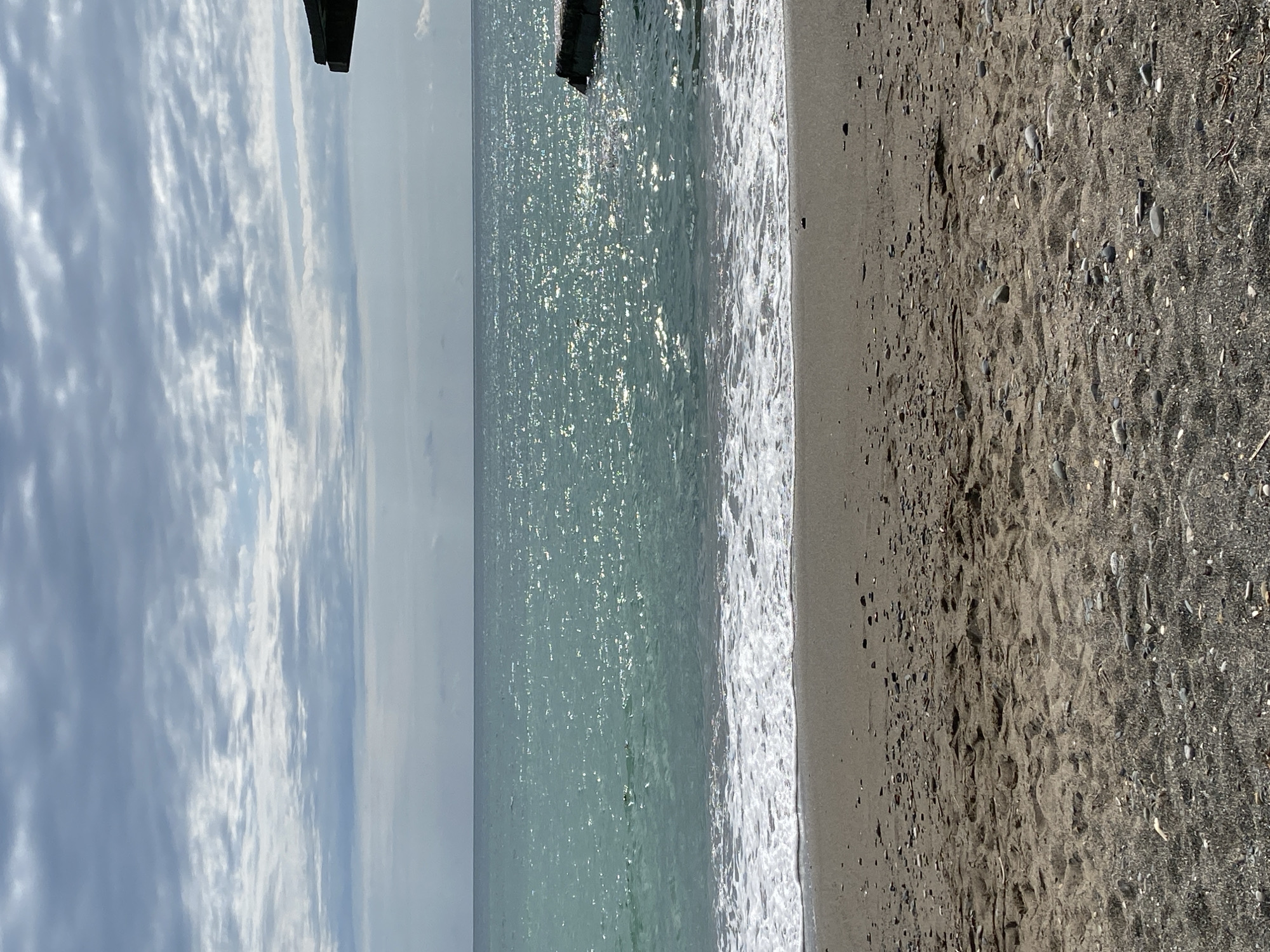 The image depicts a serene beach scene in Sokhumi, Abkhazia. The foreground is occupied by sandy beach with scattered pebbles and some debris. Gentle waves with white foam are breaking on the shore, leading to the vast expanse of the Black Sea. The water is a clear turquoise color, shimmering with sunlight. In the distance, the horizon separates the sea from a sky filled with dramatic, textured clouds. A dark, angular structure, possibly a part of a pier or a building, juts out from the upper right corner of the frame, casting a shadow on the water. A single, dark, irregularly shaped rock is visible in the sea to the right of the frame. The overall impression is one of a tranquil and overcast day at the seaside.