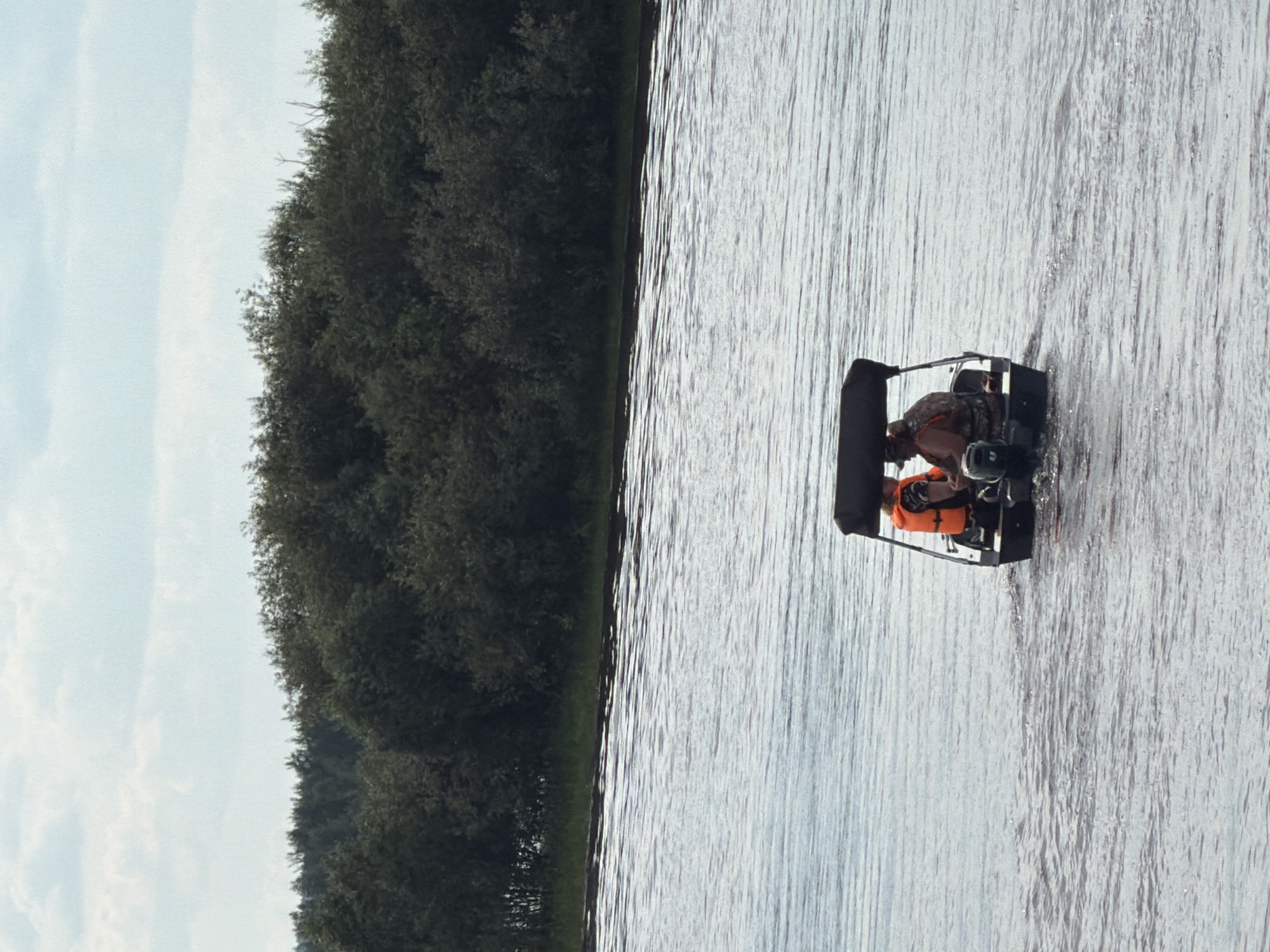 Two individuals are seated in a small motorboat moving across a body of water. The person in the front is wearing an orange life vest. The person behind them is wearing a camouflage-patterned vest. The boat has a dark canopy overhead. The water is rippled, indicating movement. In the background, a dense line of dark green trees forms the shoreline, with a pale, cloudy sky above. The scene is a natural landscape, consistent with a river or lake environment, potentially in Solikamsk, Russia, given the typical geography of the region.