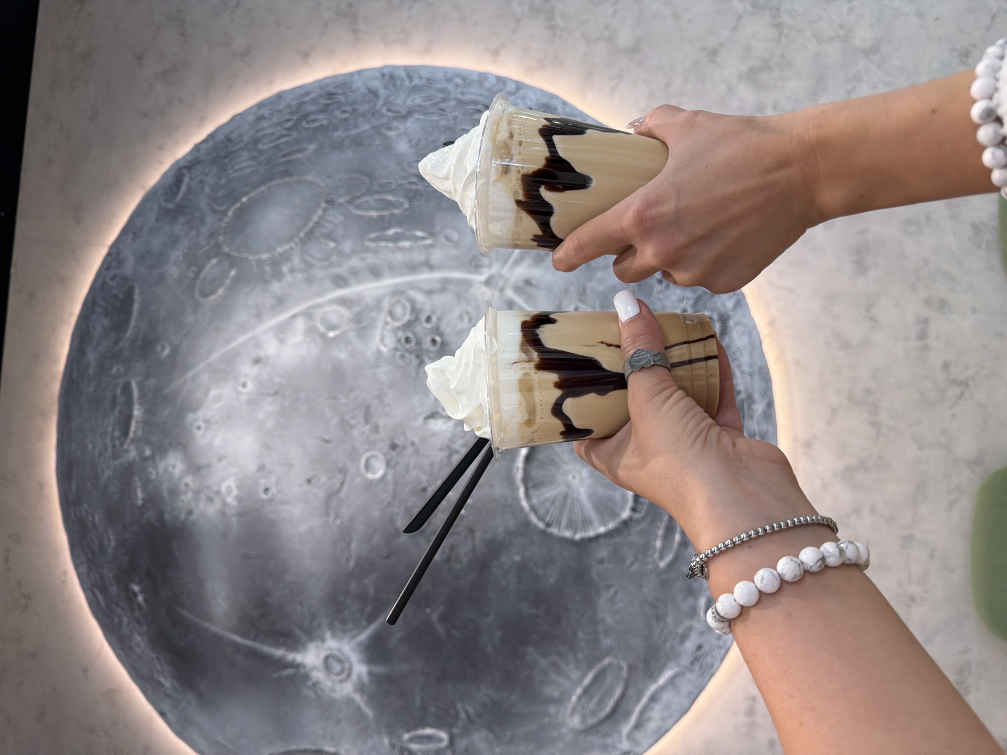 Two hands hold two iced coffee drinks in plastic cups with whipped cream and chocolate syrup drizzle. Each drink has two black straws. One hand wears a silver ring and a silver and white beaded bracelet. The other hand wears a white beaded bracelet. Behind the hands and drinks is a large, round art piece resembling the moon with craters and a lit halo. The location is Unknown, Unknown.