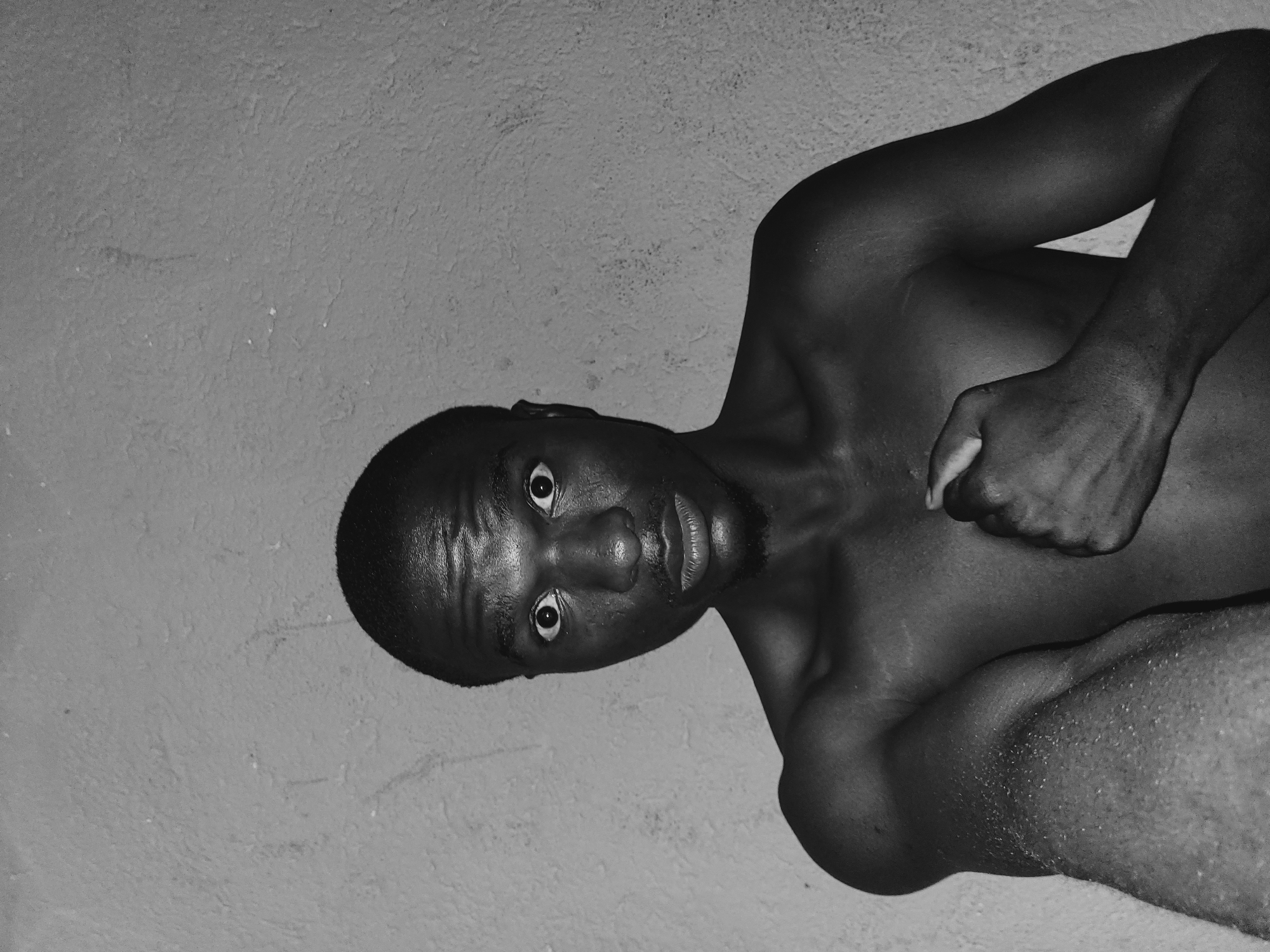A close-up, black and white photograph shows a young Black male looking directly at the camera with a surprised expression. His eyes are wide open, and his mouth is slightly parted. He is shirtless, with his left arm bent at the elbow and his hand resting on his chest. The background is a textured, gray wall. The lighting suggests an indoor setting, possibly in a residential area of Bukuru, Nigeria, based on the inferred context. The image is a portrait with no other people or objects prominently featured.