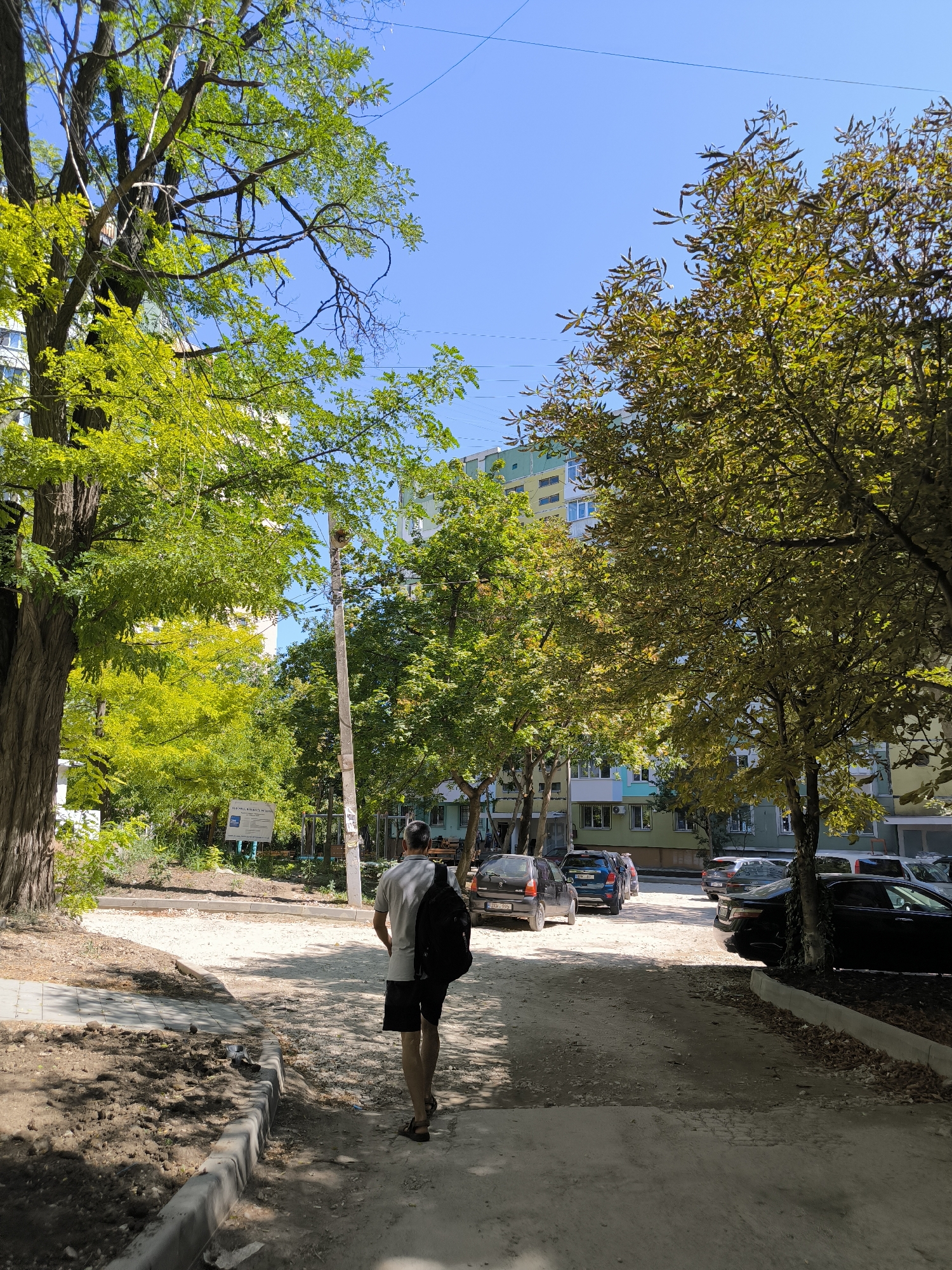 In Chișinău, Moldova, on a sunny day, a man walks away from the camera down a dusty, unpaved road lined with trees. He wears a grey t-shirt, black shorts, and sandals, carrying a black backpack over his right shoulder. Several cars are parked along the road. Buildings with colorful facades are visible behind the trees. The scene suggests a residential area with some ongoing construction or maintenance, indicated by the piles of dirt and exposed soil on the left side of the road.