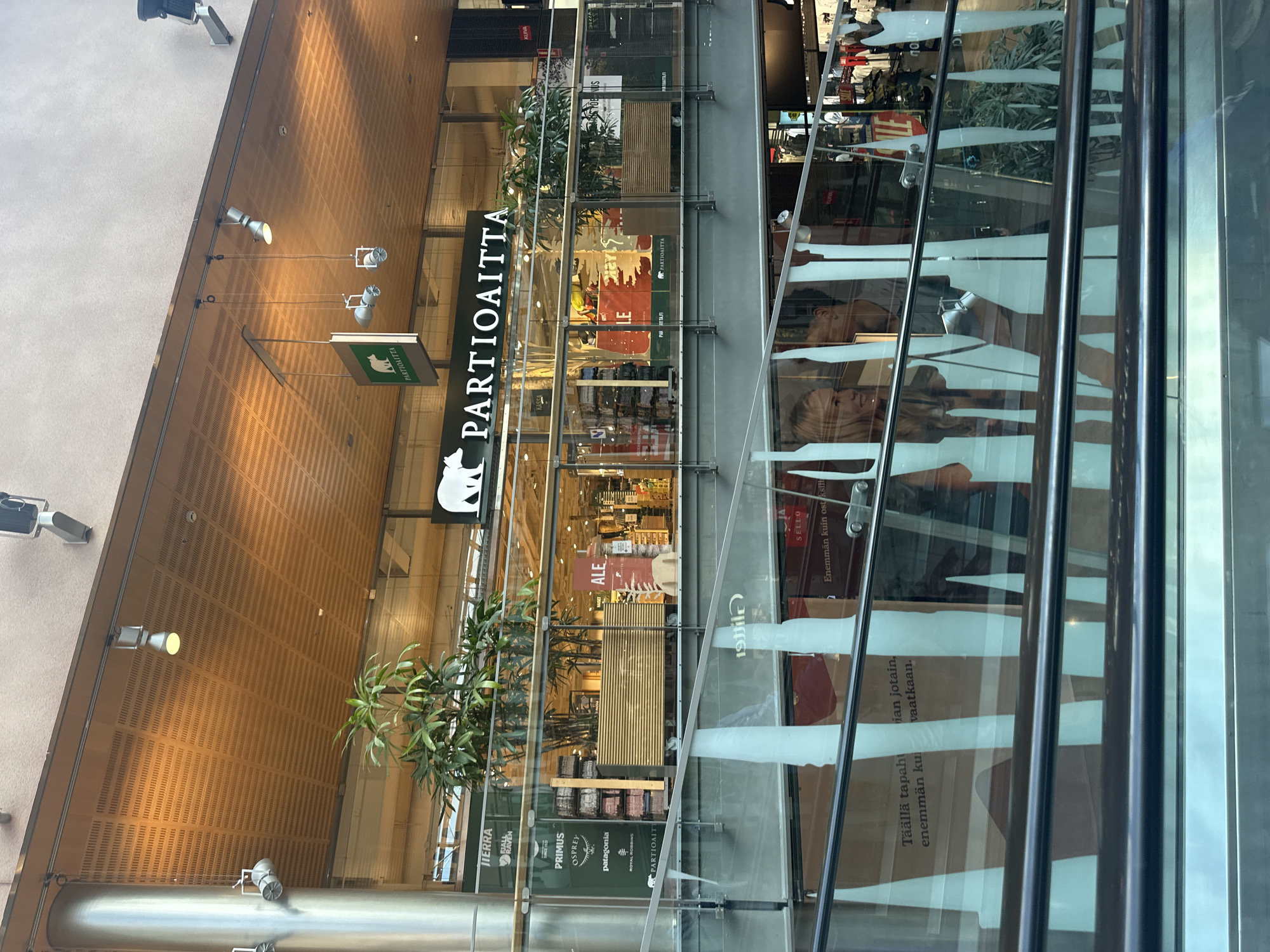 This media file shows the interior of a shopping mall, with a focus on a store called "Partioaitta". The store appears to specialize in outdoor and adventure gear, as evidenced by the logos of brands like Fjällräven, Primus, Osprey, and Patagonia visible in the image. The store's name and logo, which features a stylized animal (possibly a bear or dog), are prominently displayed on a vertical sign.
The scene is well-lit, suggesting it's daytime, and the environment appears clean and modern, typical of a commercial shopping area. Reflections on the glass storefront and railings are visible, hinting at other parts of the mall or a ceiling above. Through the glass, shelves stocked with various products and some plants can be seen. There are also some signs within the store, including one that says "ALE," indicating a sale.
The overall impression is a commercial setting, a retail store designed to attract customers interested in outdoor activities. The image is taken from a slight angle, looking into the store from the outside. There are no people clearly visible within the store itself, but there are reflections of people in the glass on the right side of the image. Text within the image includes brand names, the store's name, and promotional phrases like "Täällä tapahtuu enemmän kuin ostoksilla" which translates to "More happens here than shopping."
