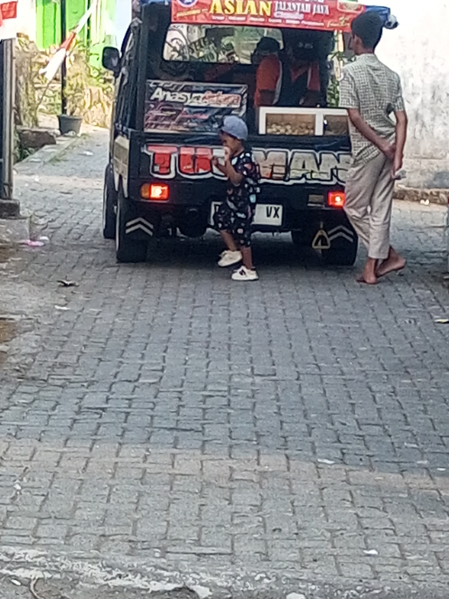 An Indonesian boy wearing a dark patterned t-shirt, dark shorts, and white sneakers is standing on a cobblestone street. He is holding a white cup and appears to be drinking from it. To his right, a man in a plaid shirt and light-colored pants is standing with his hands behind his back. Behind them is a black pickup truck with a red and white sign on top. The truck has graphics on the back. The scene is located in Andongsili, Indonesia, as indicated by the context. The environment is an outdoor urban setting with cobblestone paving.