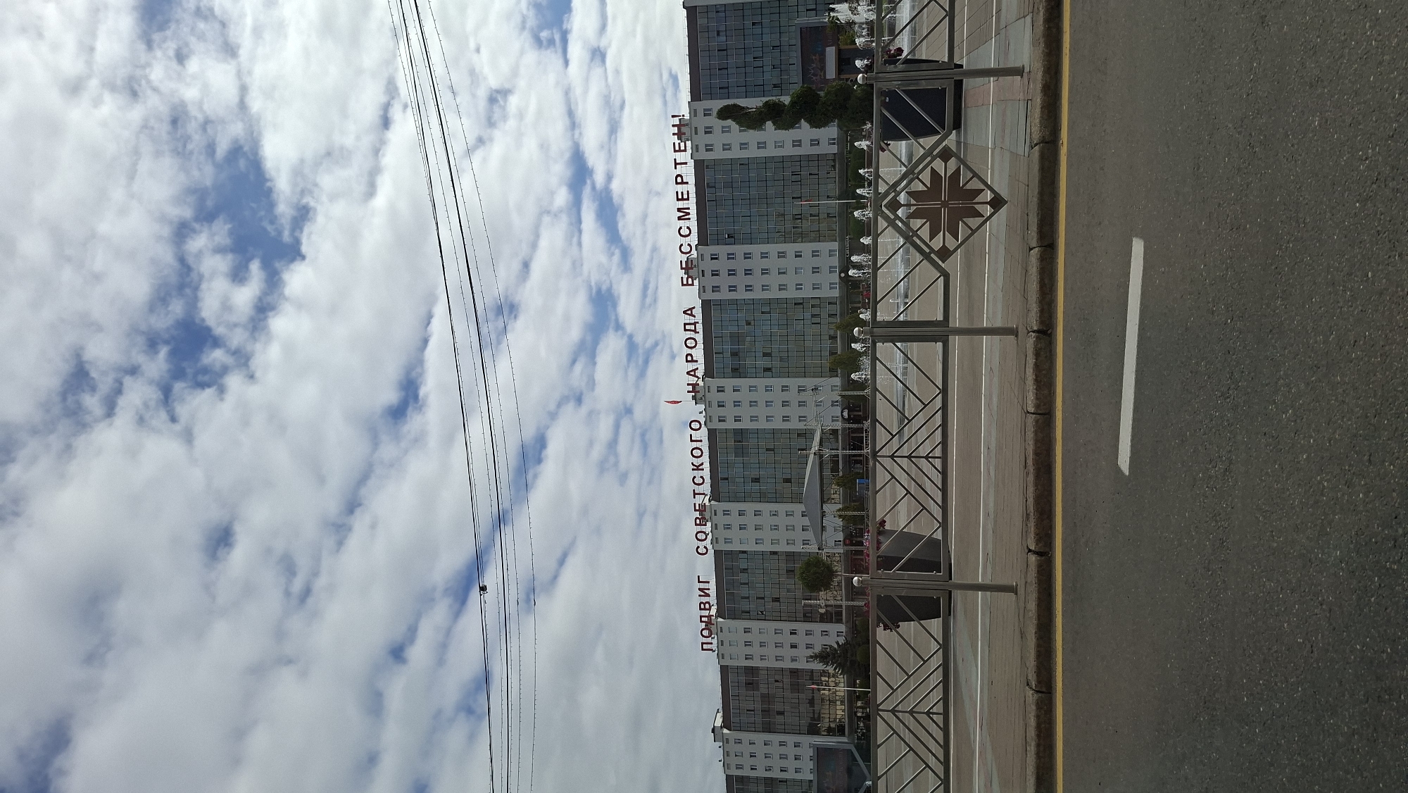 This is a medium shot, taken from a street-level perspective, of the Victory Square in Vitsebsk, Belarus. The main subject is the large, modern building that faces Victory Square. This building is characterized by its expansive glass facade and a prominent sign in red Cyrillic letters that reads "ПОДВИГ СОВЕТСКОГО НАРОДА БЕССМЕРТЕН!". This translates to "The feat of the Soviet people is immortal!". A small red flag is visible atop the building.
In front of the building, a fountain is active, with several jets of water visible. The square area is delineated by a decorative metal fence with a geometric pattern. On the fence, there are two dark planters with greenery and some pink flowers. To the right of the fence, a tall, spiral-shaped evergreen tree stands.
The foreground of the image shows a paved road with a yellow line and a white horizontal line marking. The sky is partly cloudy with a mix of white clouds and patches of blue sky. The overall impression is of a sunny day. No people are clearly visible in the image, though there are some indistinct figures in the distance near the fountain. The scene appears to be a public space, likely a tourist or civic landmark.