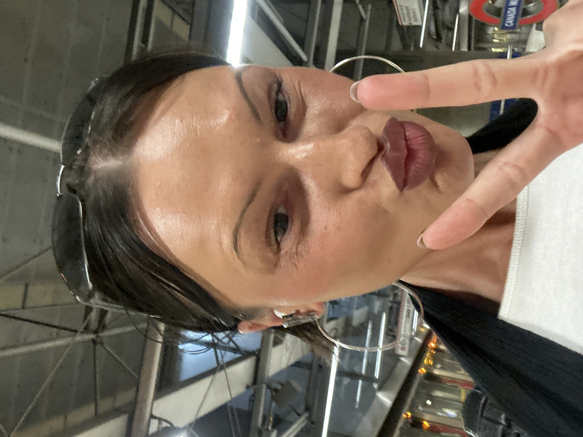 A rotated, close-up portrait features a woman posing playfully indoors. She is looking towards the camera with a kissy face, her lips adorned with dark lipstick. Her right hand is raised, making a peace sign near her face. She has dark hair, styled back, with sunglasses pushed up onto her head and large hoop earrings. She appears to be wearing a white top and a dark, ribbed garment underneath. The background indicates an indoor public or transport setting, characterized by industrial-style ceiling structures, metal beams, and artificial lighting. A significant detail in the background is a partially visible red circular sign, identifiable as a London Underground (Tube) roundel, with the text "CANADA W" clearly legible, indicating "Canada Water" station. This confirms the location as London, United Kingdom. The image captures a candid and cheerful moment.