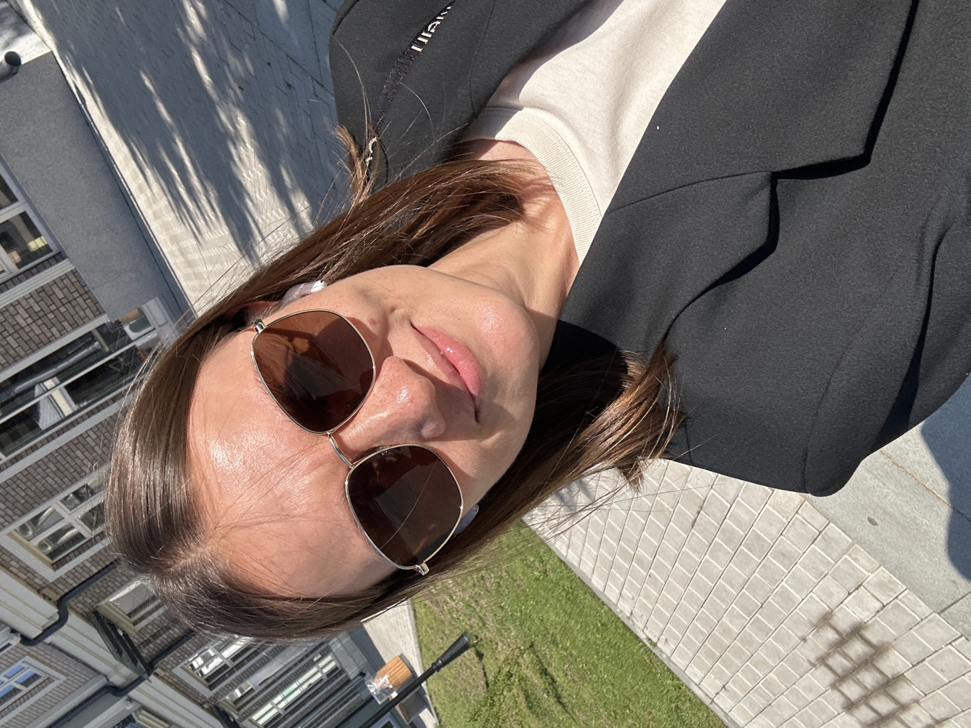 A woman with brown hair is taking a selfie outdoors, wearing a black blazer, a light-colored t-shirt, and sunglasses. She has earbuds in. The background shows a building with brickwork and windows, a paved walkway with a grass area to the left, and trees casting shadows on the ground. The scene is lit by sunlight. The location is likely outside a residential building in the city of Kazan, Russia, given the architectural style and typical landscaping.